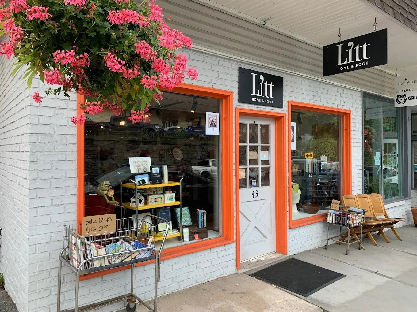 Exterior view of a bookstore named 'Litt Home & Book' with white brick walls and orange window frames. There are shopping carts and a bench outside, and a large pink flower plant hanging above the entrance.