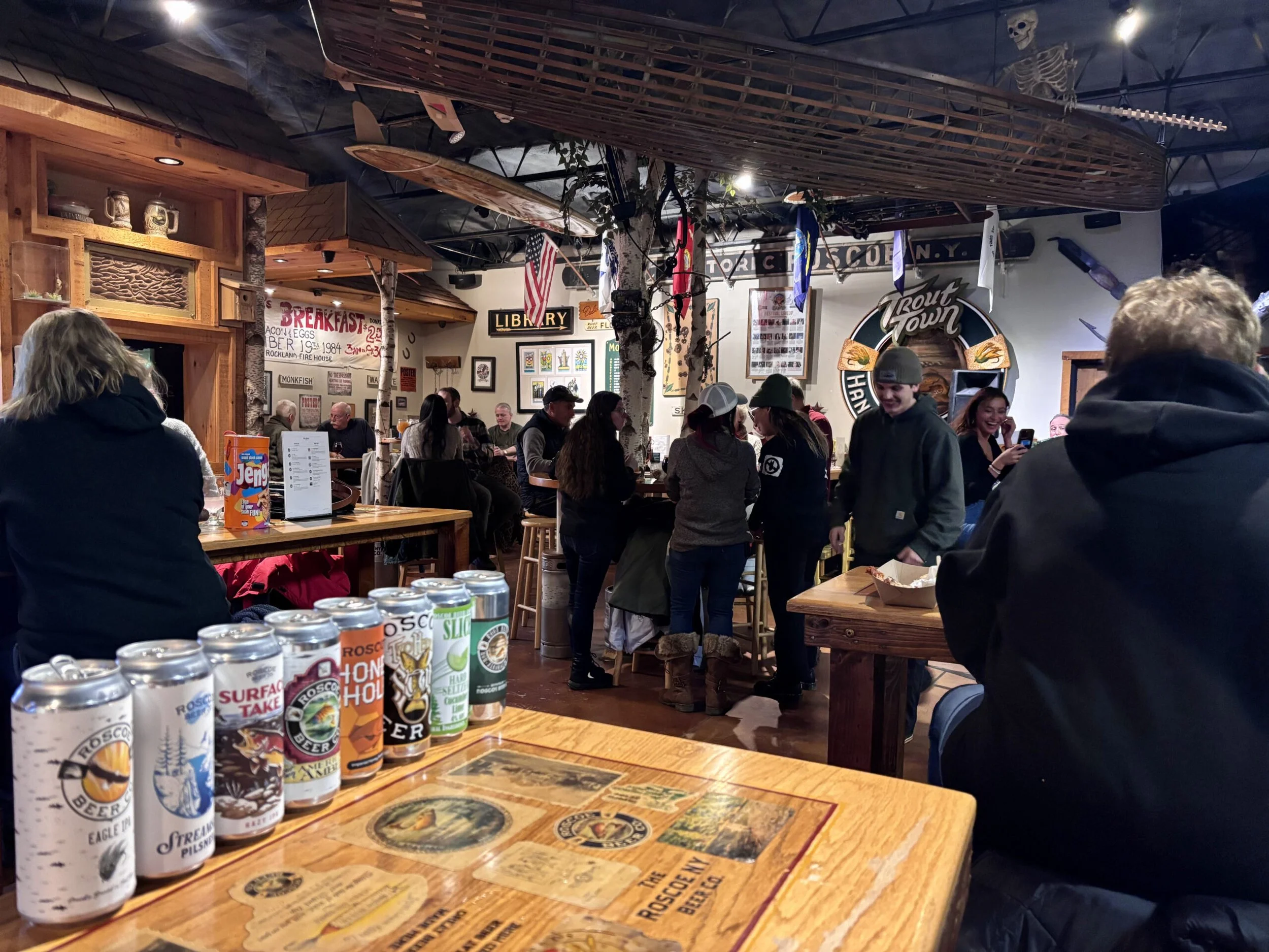 Inside a lively bar or brewery with wooden decor, people sitting and standing, some wearing winter clothing, colorful beer cans on the table in the foreground, and various signs and decorations on the walls.