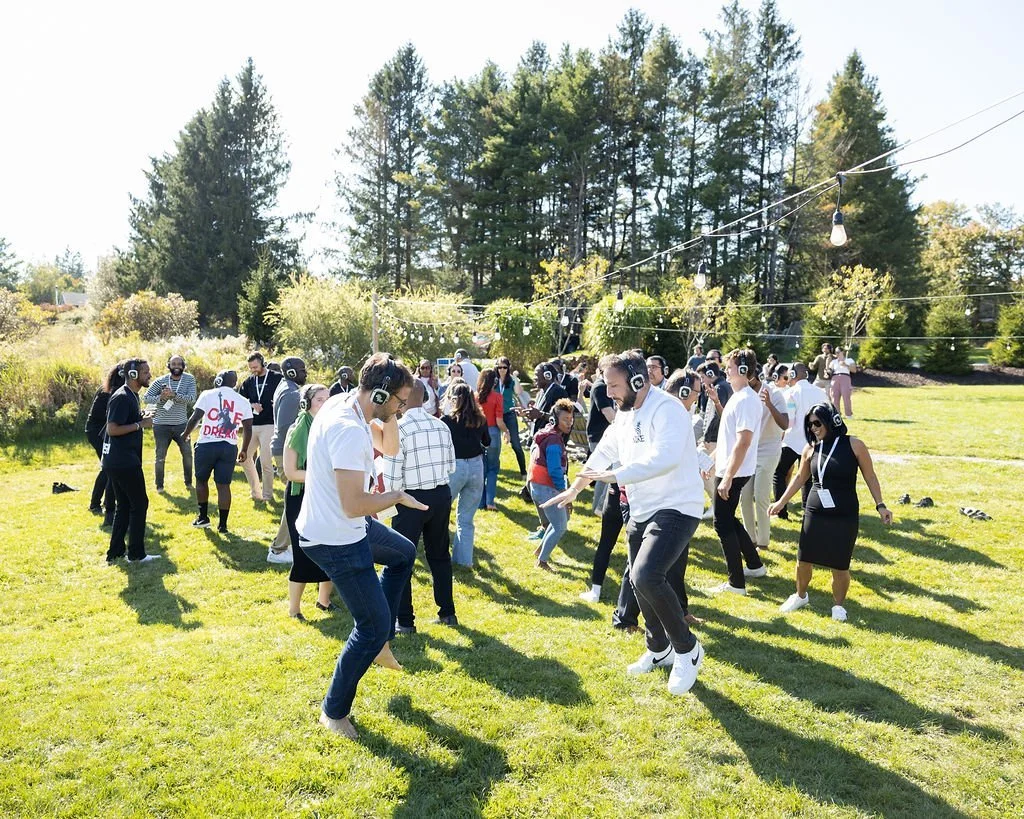 People dancing and listening to music with headphones outdoors on a sunny day, in a grassy field with trees in the background.