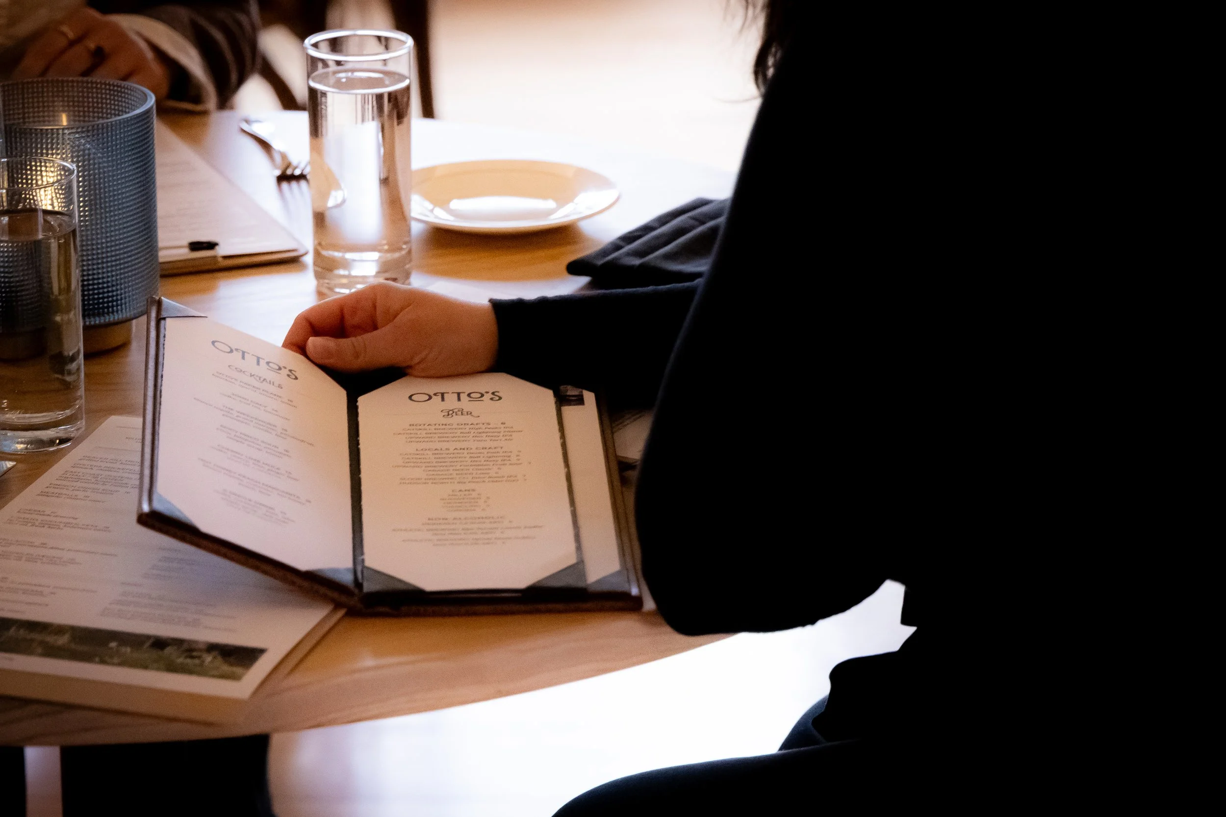 Person sitting at a wooden table in a restaurant, looking at a menu. The table has a tall glass of water, a smaller glass, a notepad, and a plate. The image shows the person's hand holding the menu and part of their arm and torso, with other diners and tables visible in the background.