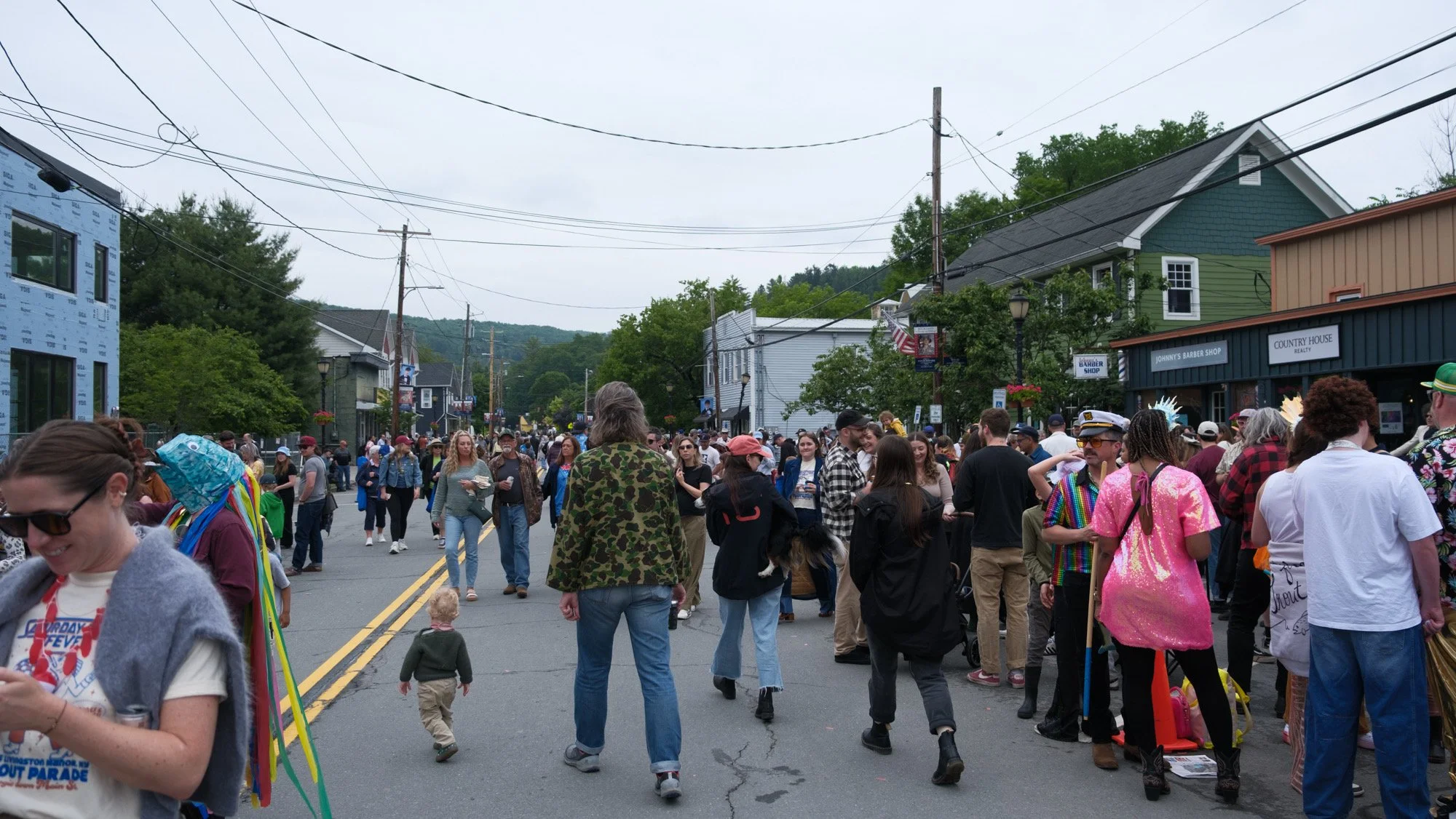 Crowd of people walking on a city street during a festival or parade, with colorful buildings and storefronts on the sides and power lines overhead.