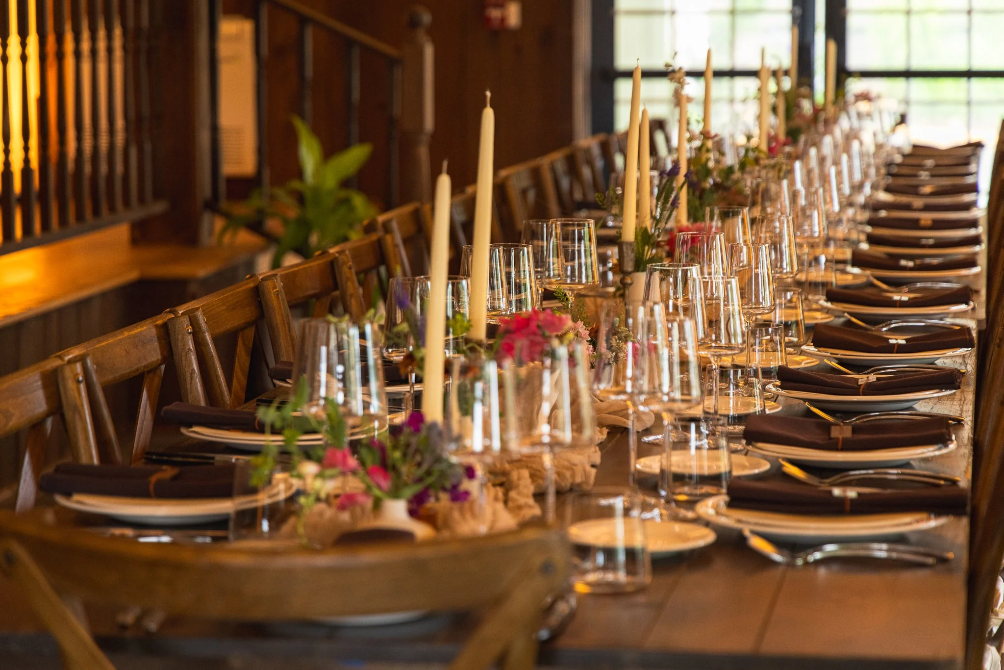 A long banquet table set with plates, silverware, glasses, and black napkins, decorated with flowers and candles in a warmly lit room.