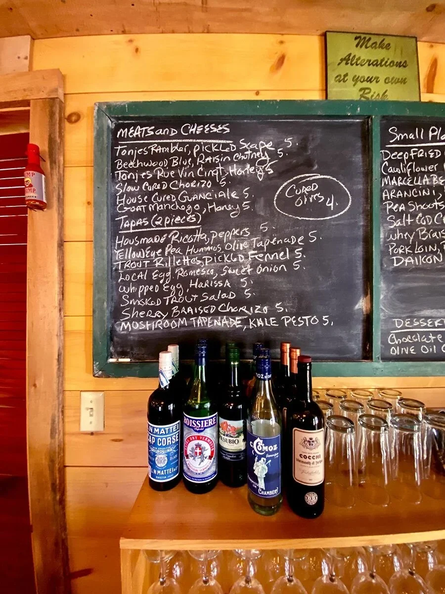 A chalkboard menu listing meats, cheeses, and small plates in a restaurant. Bottles of wine and glasses on a wooden counter in front of the menu.