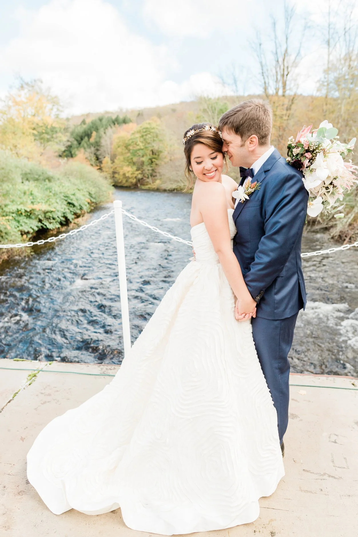 A bride and groom standing close, holding hands, smiling, near a river with trees showing fall colors in the background.