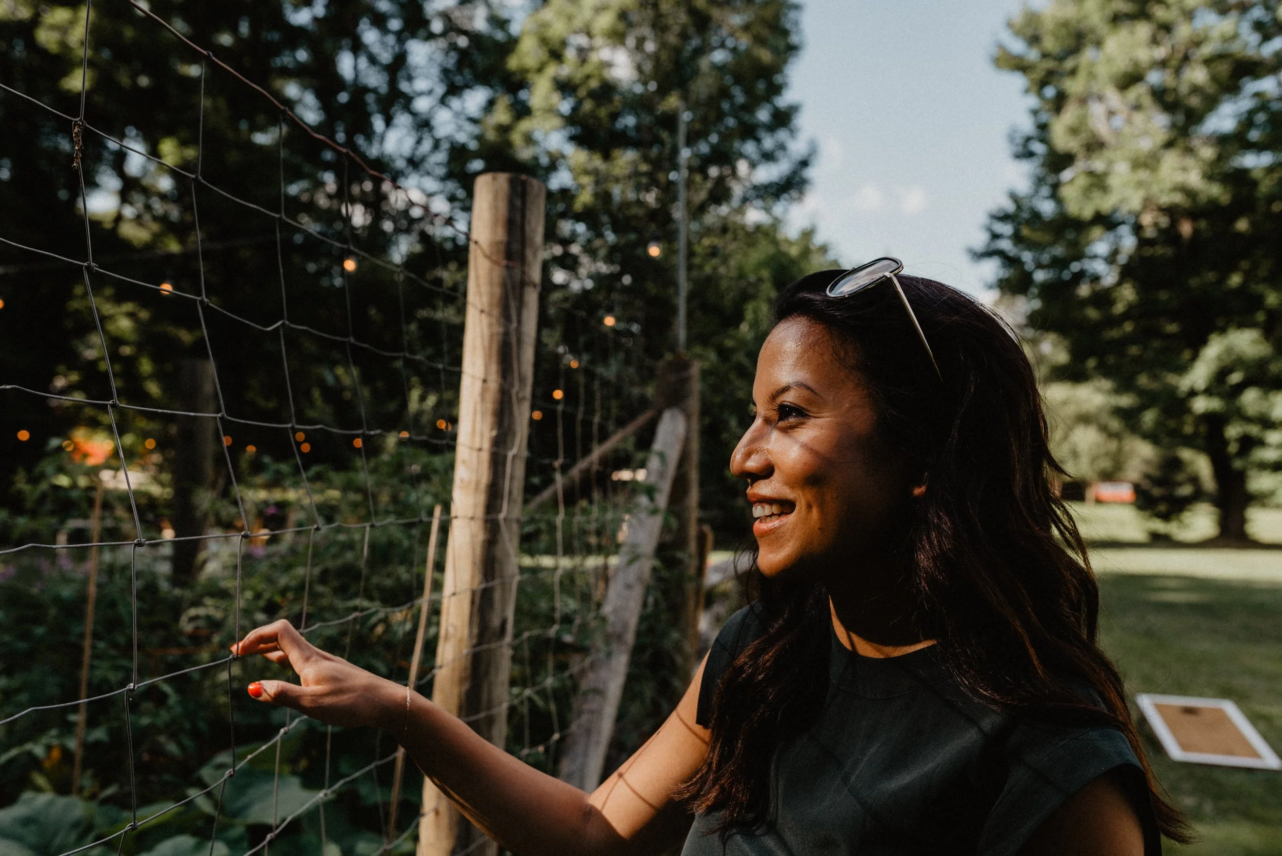 A woman with dark hair and glasses on her head smiling and reaching out to a wire fence in an outdoor setting with trees and string lights.