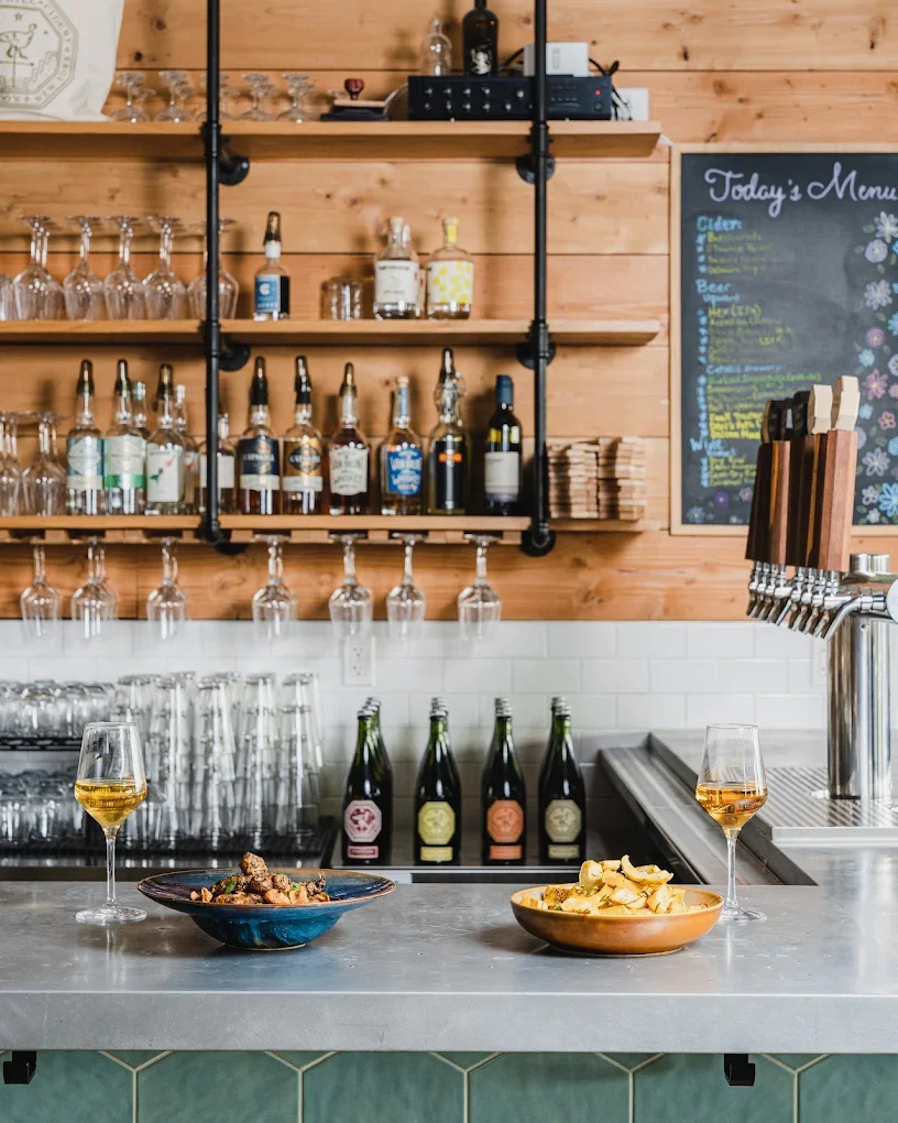 Bar counter with two glasses of white wine, bowls of snacks, and bottles of beverages on a gray countertop. Shelves with alcoholic bottles and glassware are in the background, along with a chalkboard menu on a wooden wall.