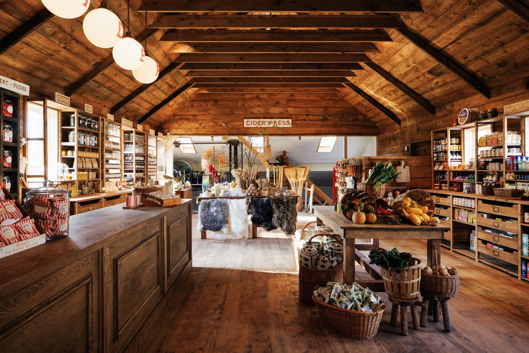 Interior of a rustic store with wooden walls and ceiling, displaying shelves of jars, bags, and canned goods, with a central table holding produce and decorative items, illuminated by warm lighting.