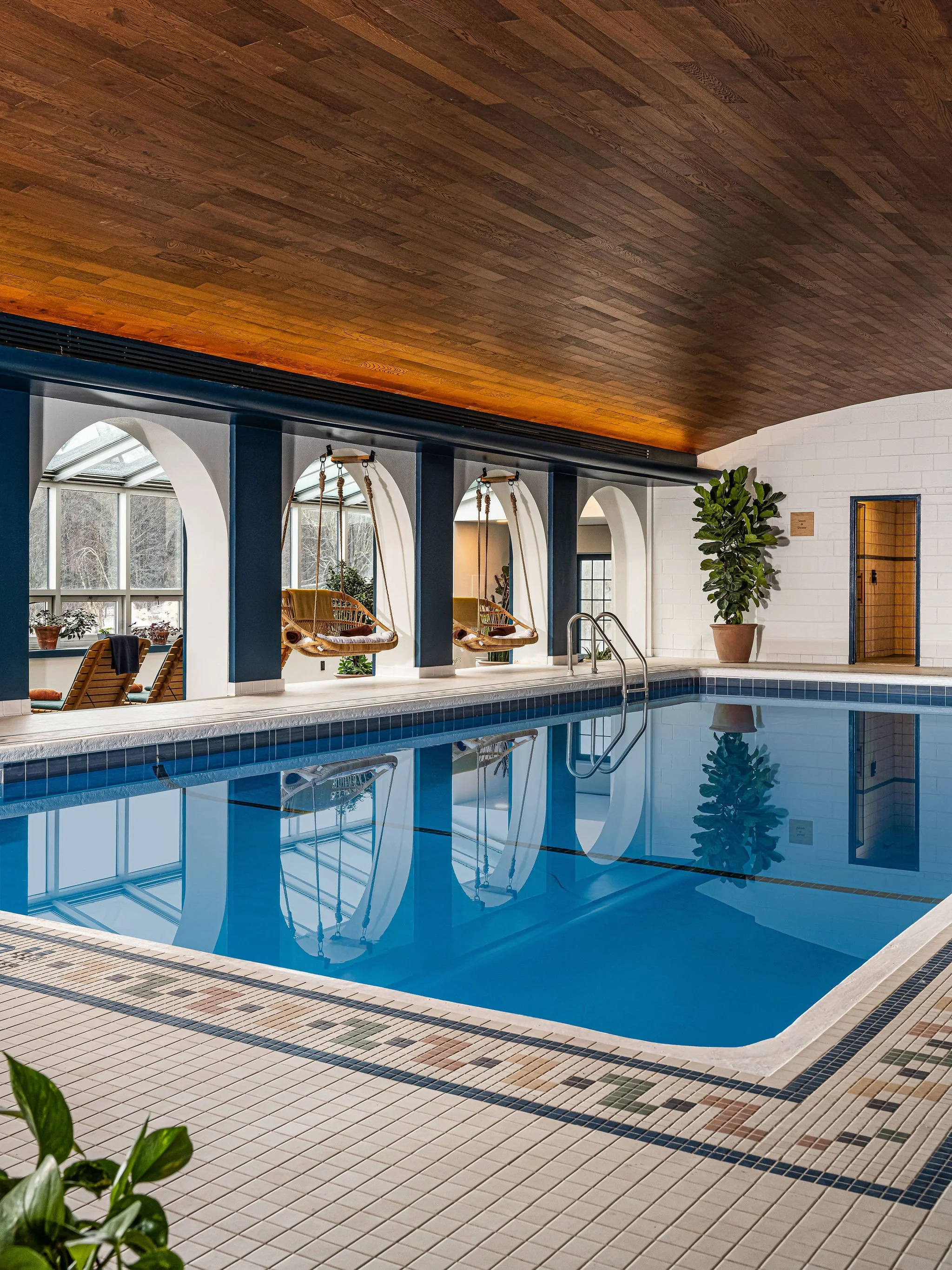 Indoor swimming pool with blue tiles, surrounded by a tiled deck, with wooden ceiling, large arched windows, and a potted plant.