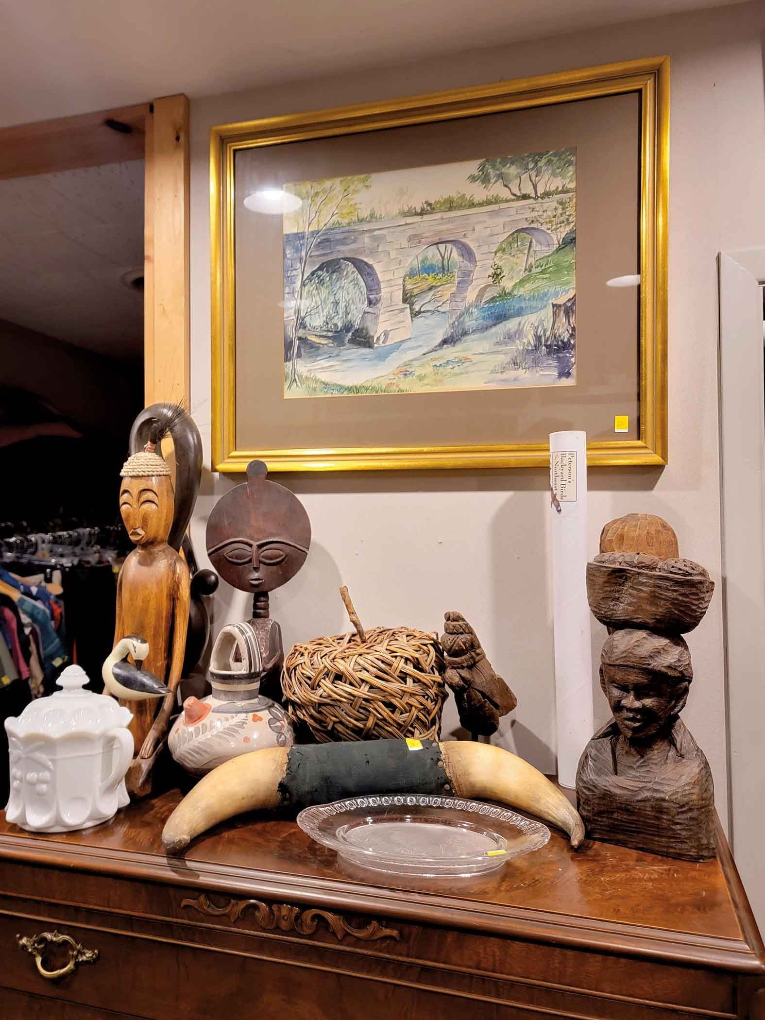 Assorted decorative items including tribal sculptures, a woven basket, a horn, a ceramic dish, and a teapot, arranged on a wooden table below a framed landscape painting with arches over a river.