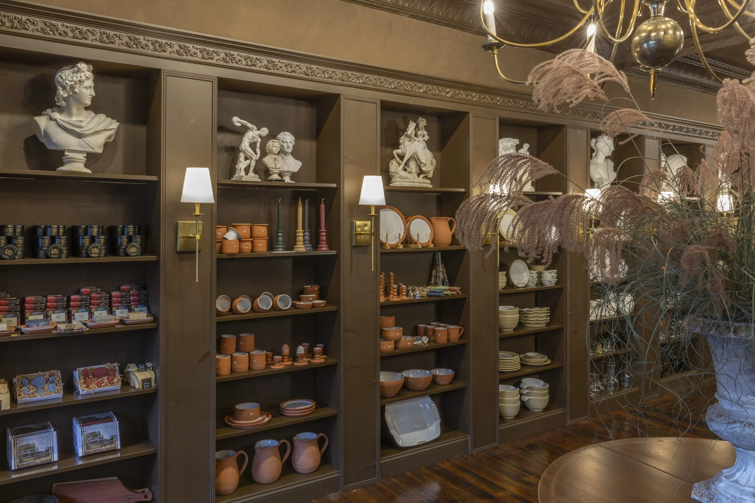 Wooden shelves in a store display vintage bust sculptures, pottery, and decorative items with a large floral arrangement in a white vase in the foreground.