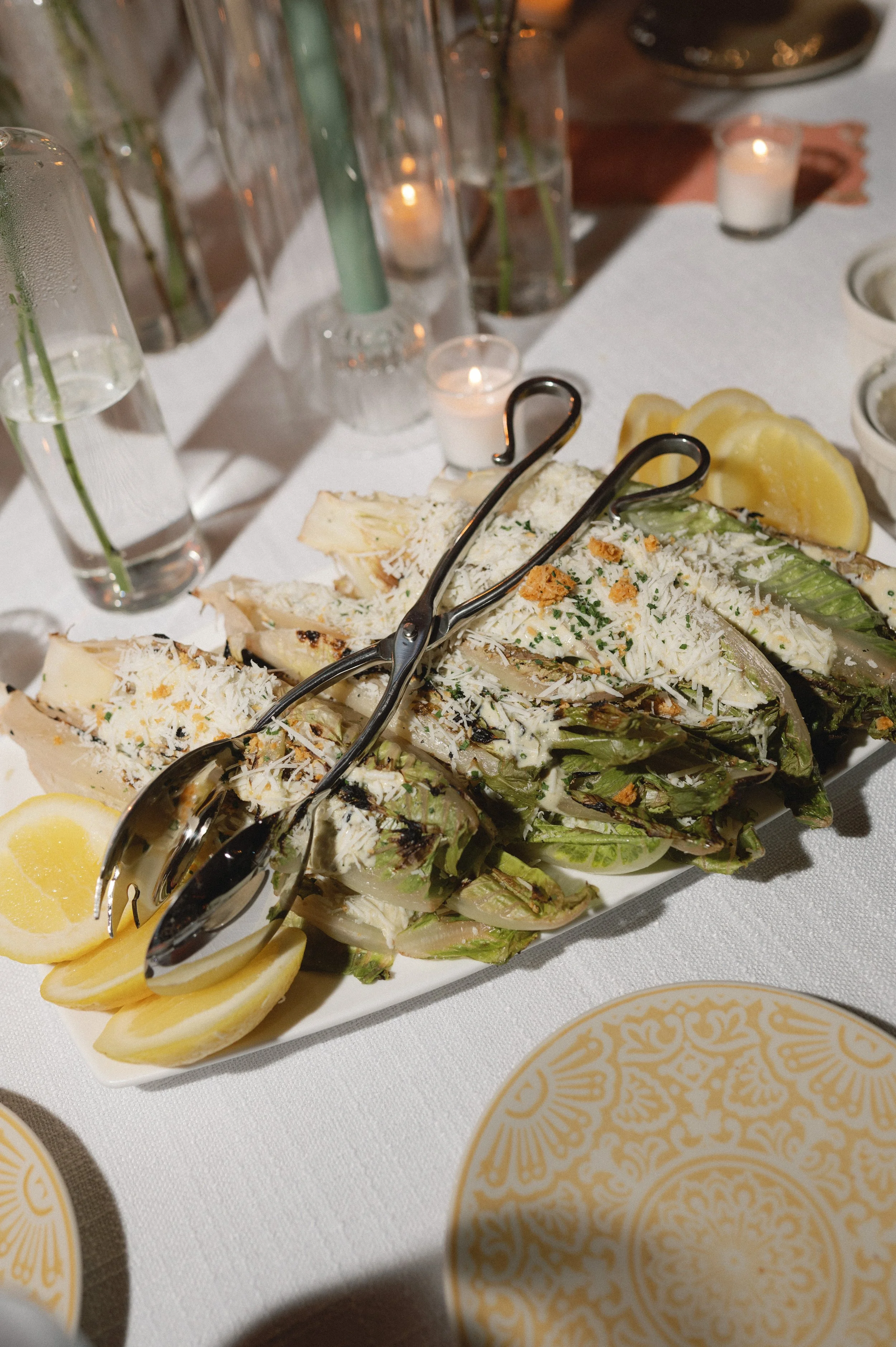 Grilled Caesar salad wedges topped with cheese, served with lemon wedges on a white platter, surrounded by candles and tableware.