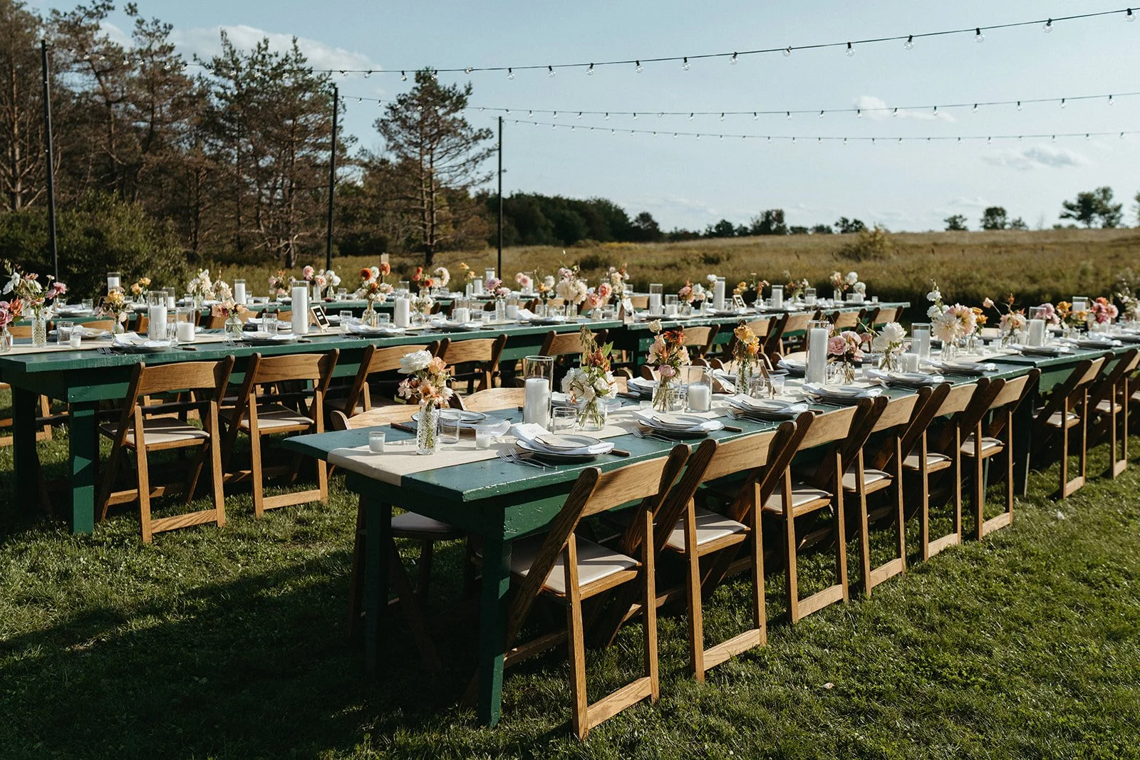 Long outdoor dining table set with flowers, candles, and dinnerware on a grassy field under string lights, with trees and open sky in the background.