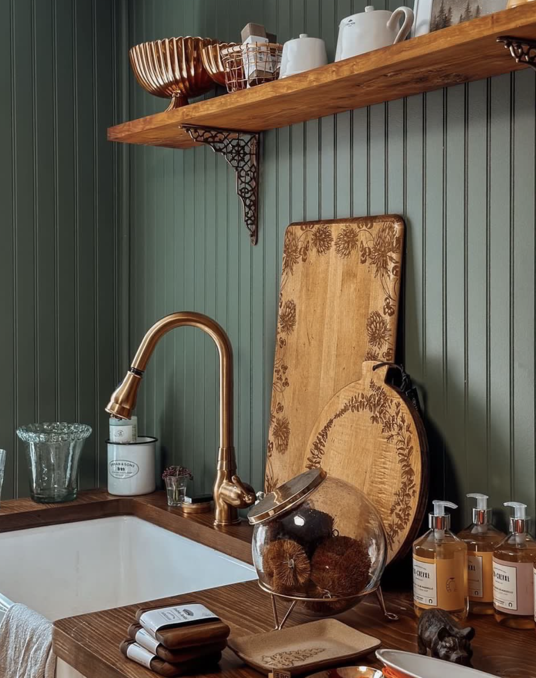 Kitchen sink with a bronze faucet, wooden cutting boards with floral designs, glass jar with brushes, and bottles of soap on a wooden countertop, green paneled wall, wooden shelf with dishes and kitchenware.