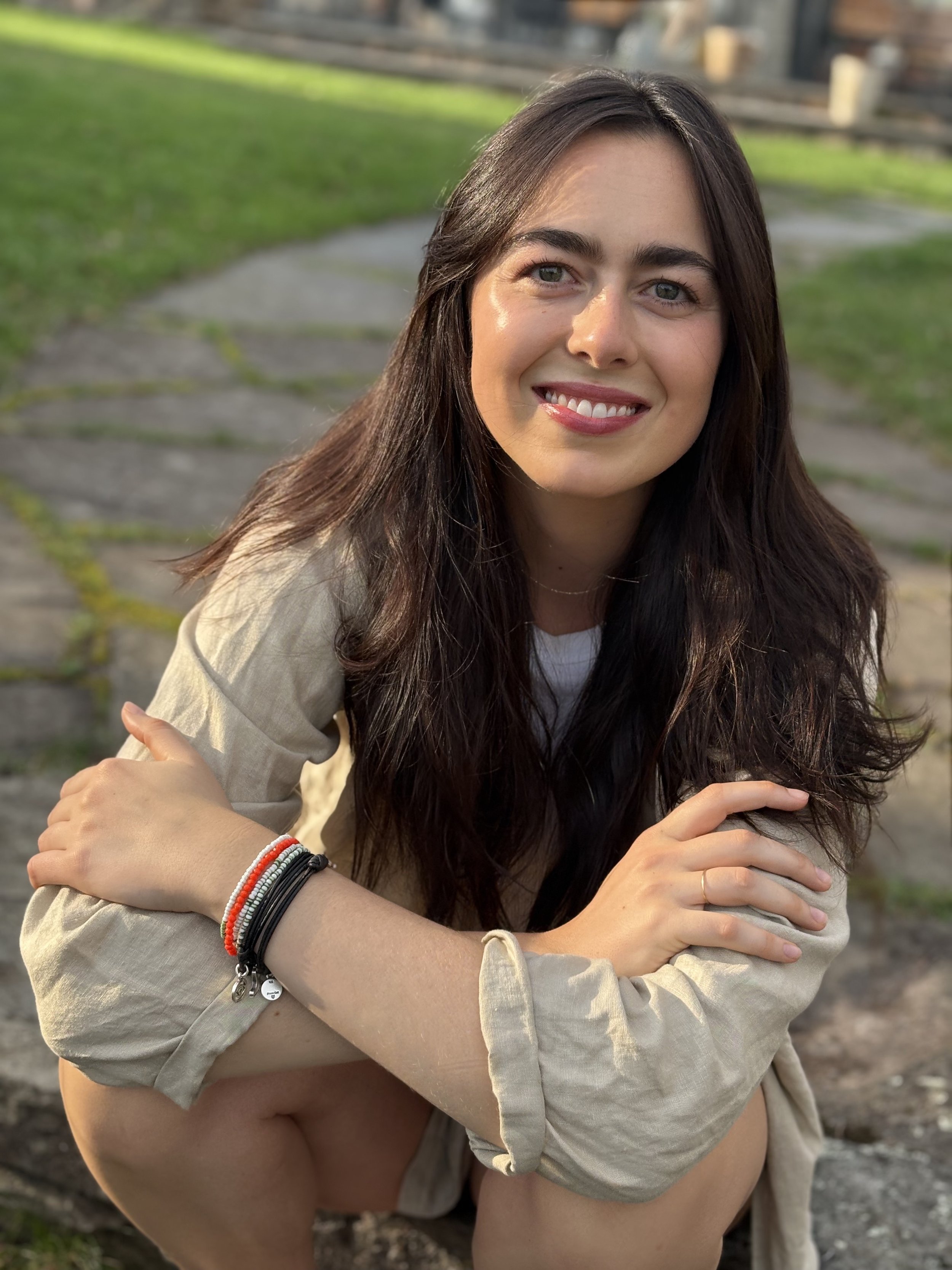 A woman with shoulder-length dark hair and a denim jacket smiling outdoors in a park or garden with white flowers and trees, people sitting at tables, and tall buildings in the background.