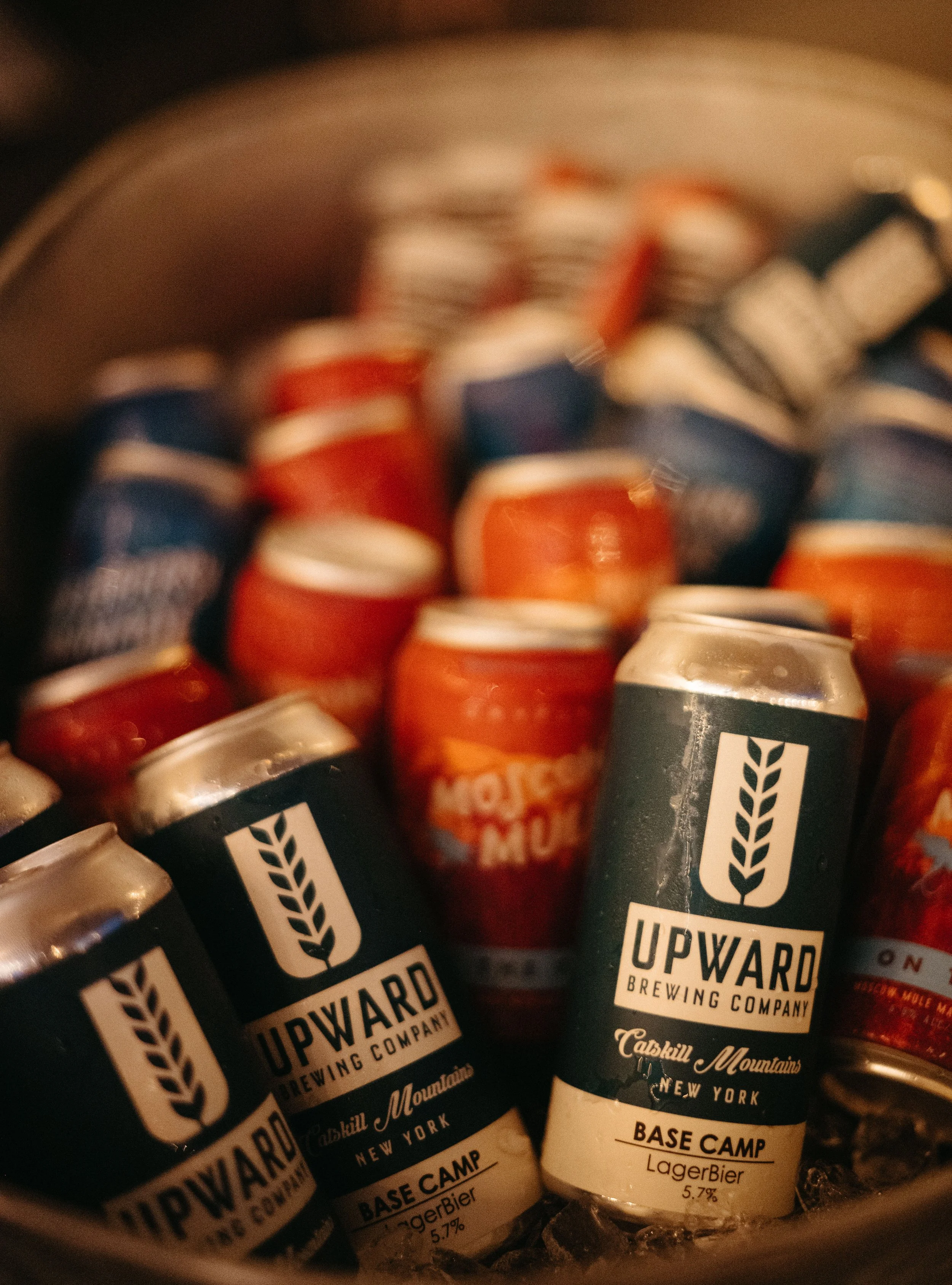 Cans of Upward Brewing Company beer, labeled 'Base Camp LagerBier 5.7%', inside a cooler with ice, with other cans in the background.