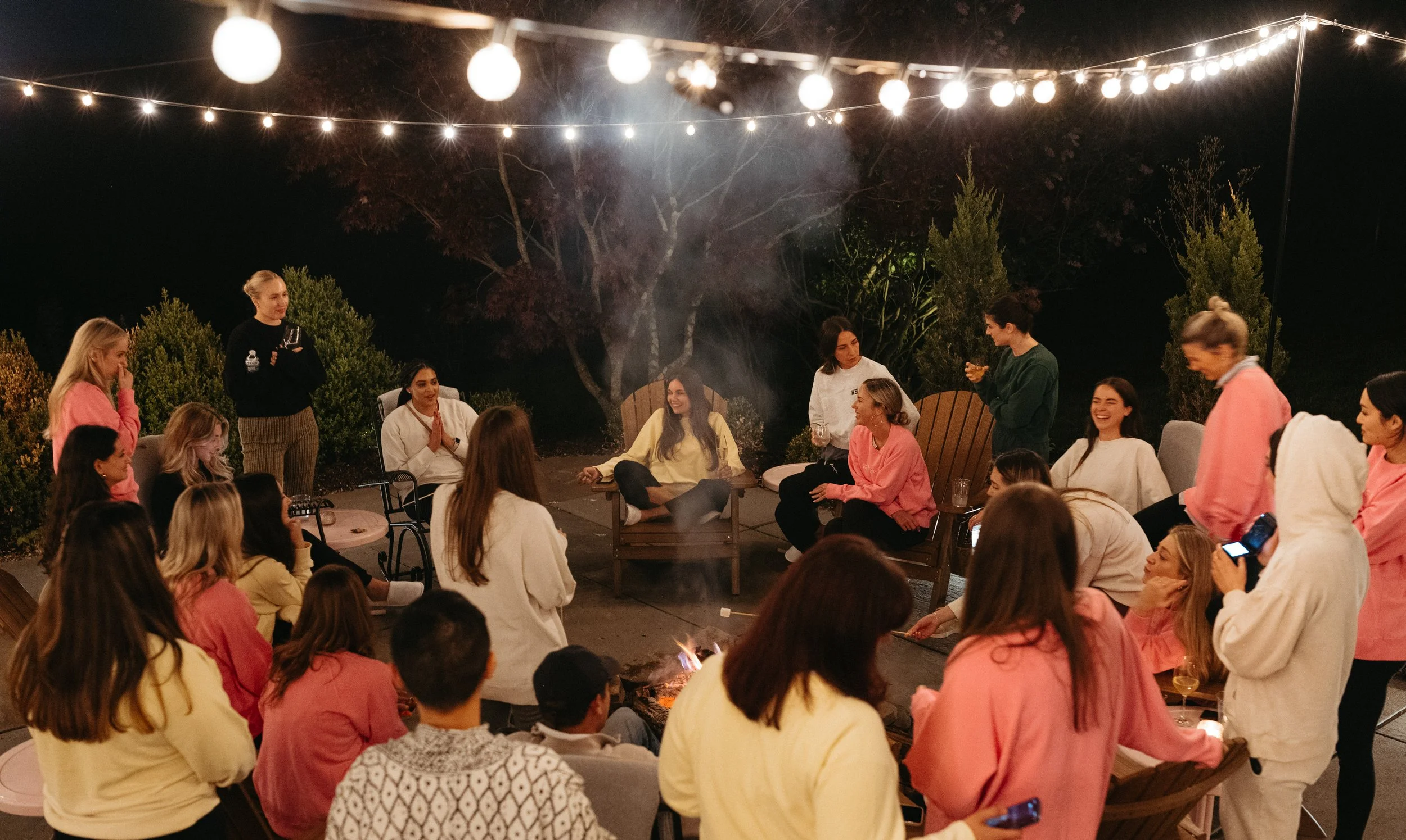 A nighttime outdoor gathering of people, mostly women, sitting and standing around a fire pit. String lights hang above, illuminating the scene. People are chatting and smiling in a relaxed atmosphere.