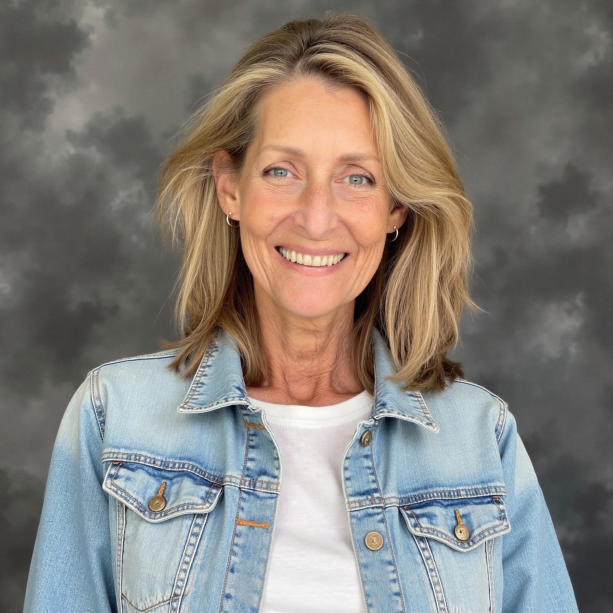 A woman with shoulder-length brown hair, smiling, wearing a gray sweater over a white collared shirt, standing in front of a wooden wall.