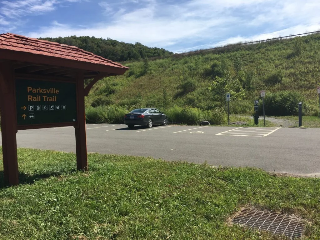 Parking lot at Parksville Rail Trail with a parked car, designated parking spaces, a sign indicating the trail, and a grassy hillside in the background.
