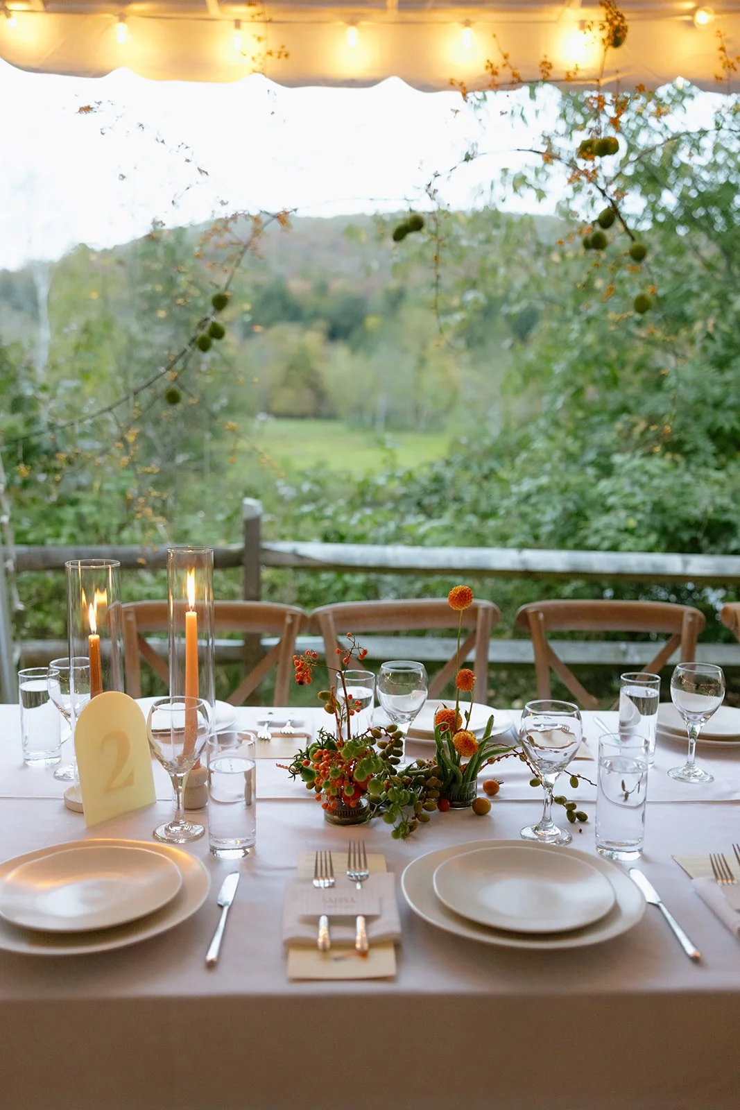 A beautifully set outdoor dining table with white plates, wine glasses, and cutlery, decorated with a floral centerpiece of orange and green blossoms, lit by candles in tall glass holders, overlooking a lush green landscape with trees and hills.