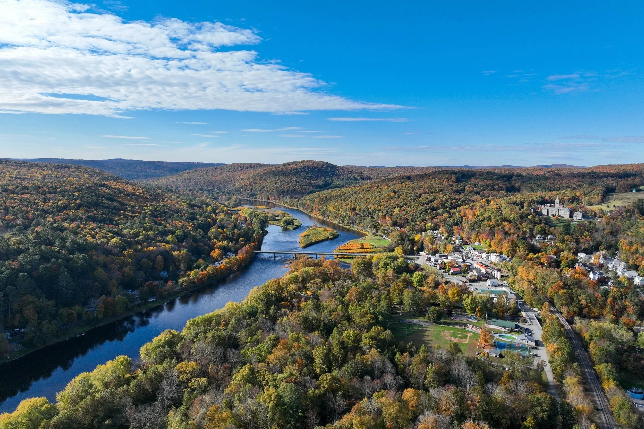 Aerial view of a river winding through a forested valley with a small town nearby under a partly cloudy blue sky.
