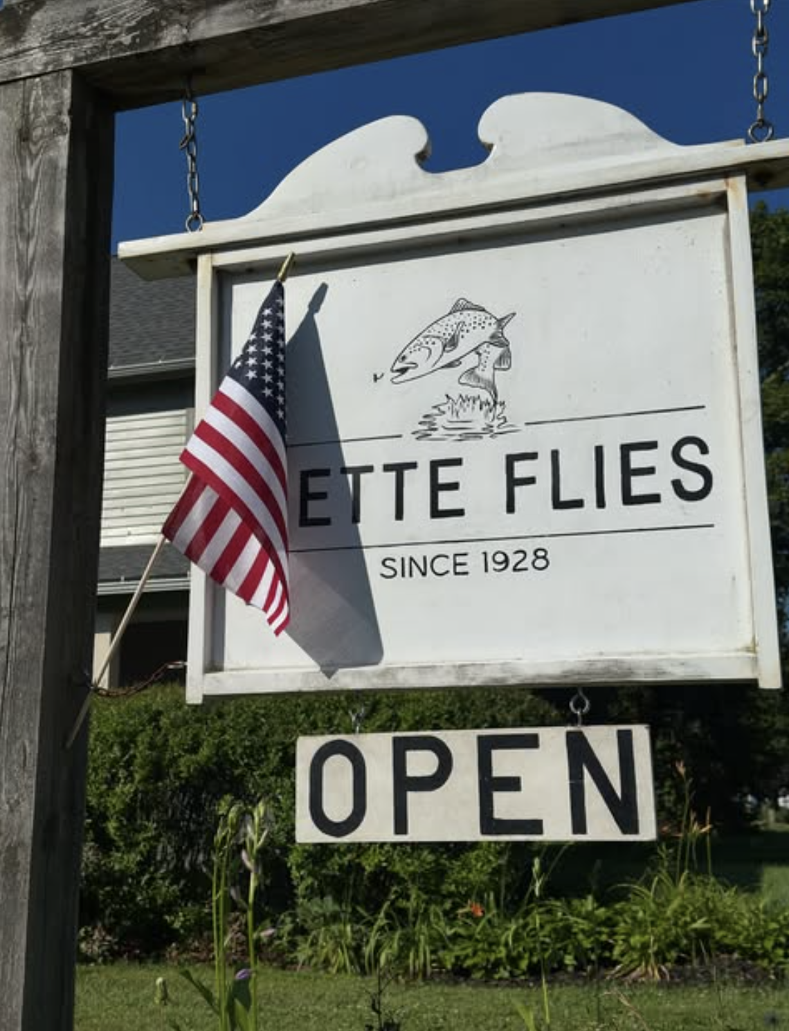 A white sign with black writing that reads 'Ette Flies Since 1928,' with an illustration of a fish jumping out of water. An American flag is attached to the sign. Below, there is a smaller sign with the word 'OPEN' in black letters. The sign is mounted on a wooden frame outdoors with some greenery and a house in the background.