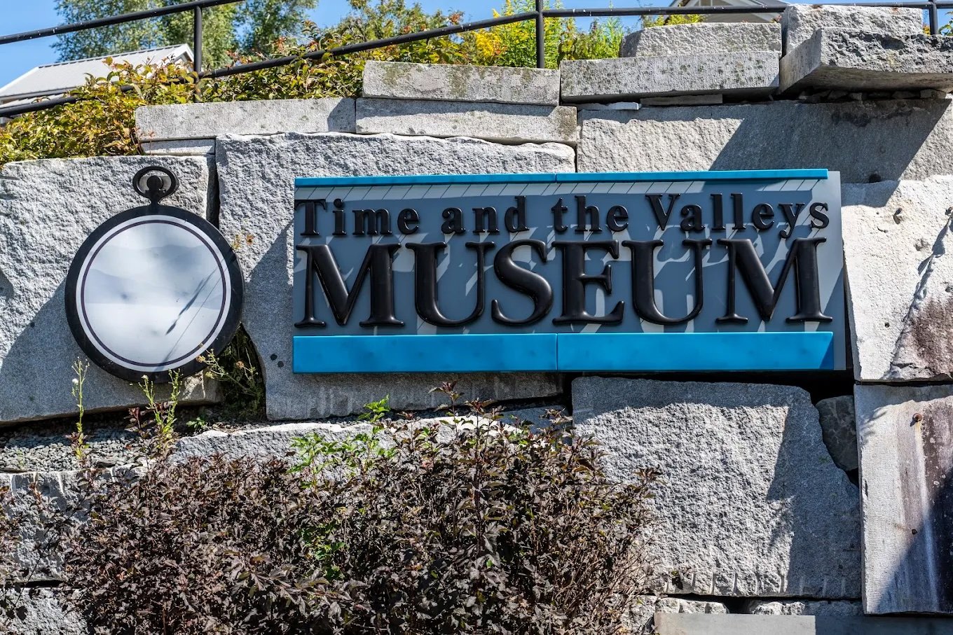 Sign for the Time and the Valleys Museum mounted on a stone wall, with a decorative clock face to the left, outdoors during daytime.