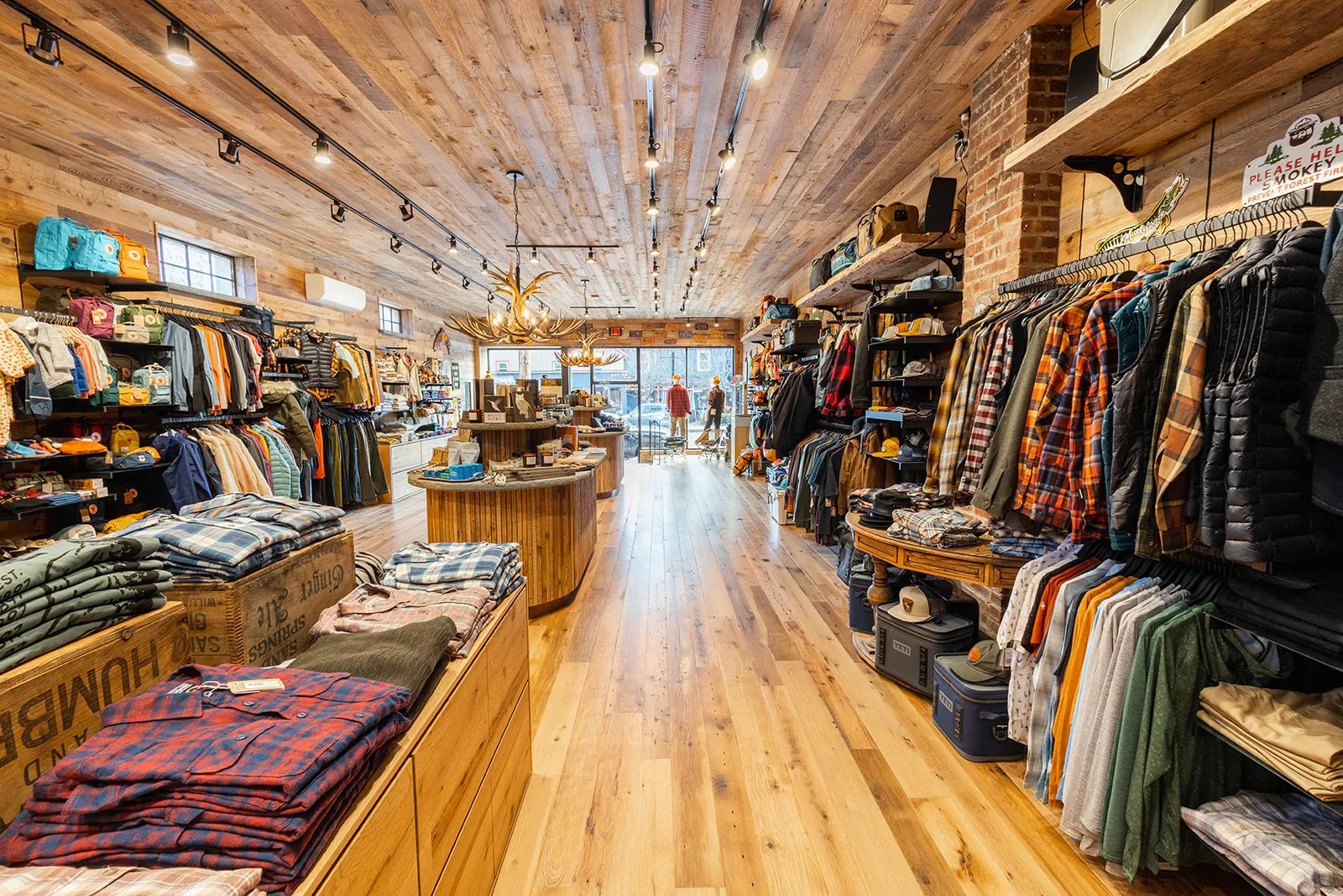 Interior of a clothing store with wooden floors and ceiling, displaying racks of clothing on both sides, mannequin standoff near the large front window, and rustic decor including antler chandelier and brick wall accents.