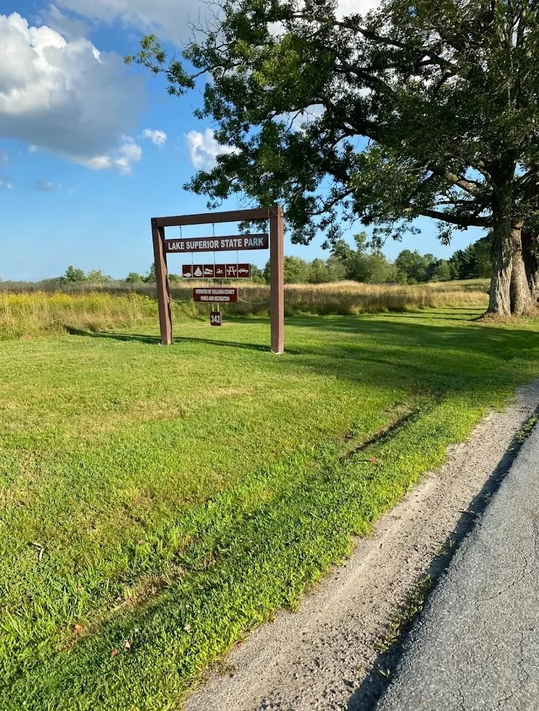 Signboard for Lake Superior State Park beside a grassy area and a large tree, with a partly cloudy sky in the background.