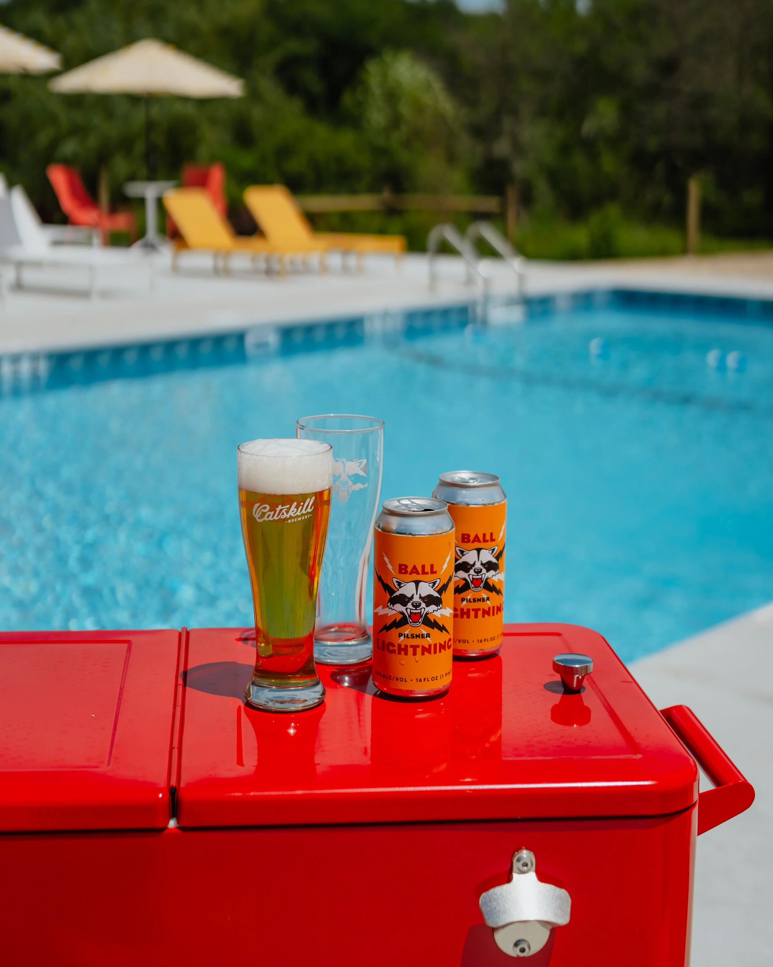 A red outdoor cooler with a glass of beer, two cans of Ball Lightning beer, and a glass of water on top, near a swimming pool with lounge chairs and umbrellas in the background.