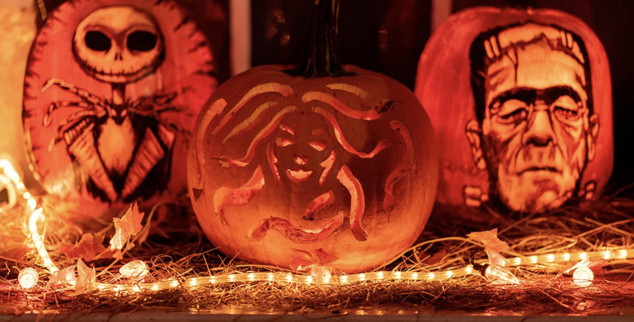 Decorative Halloween pumpkins with carved faces and designs, illuminated from within, surrounded by string lights and autumn leaves.