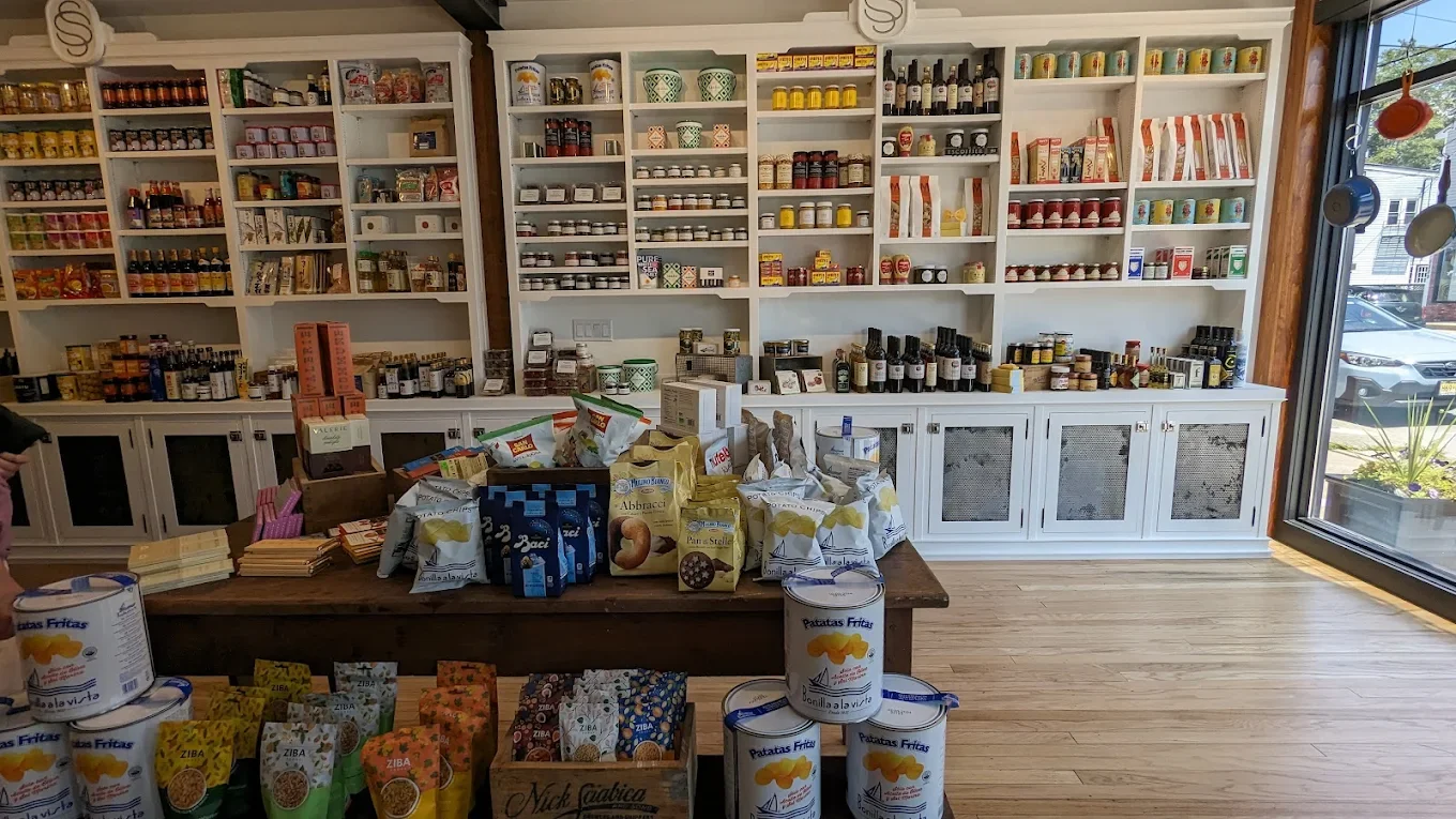 Inside a store with white shelves filled with jars, cans, and packaged goods, and a large wooden table displaying assorted grocery items, including cans of potato chips and snack packages. There is a large window to the right.