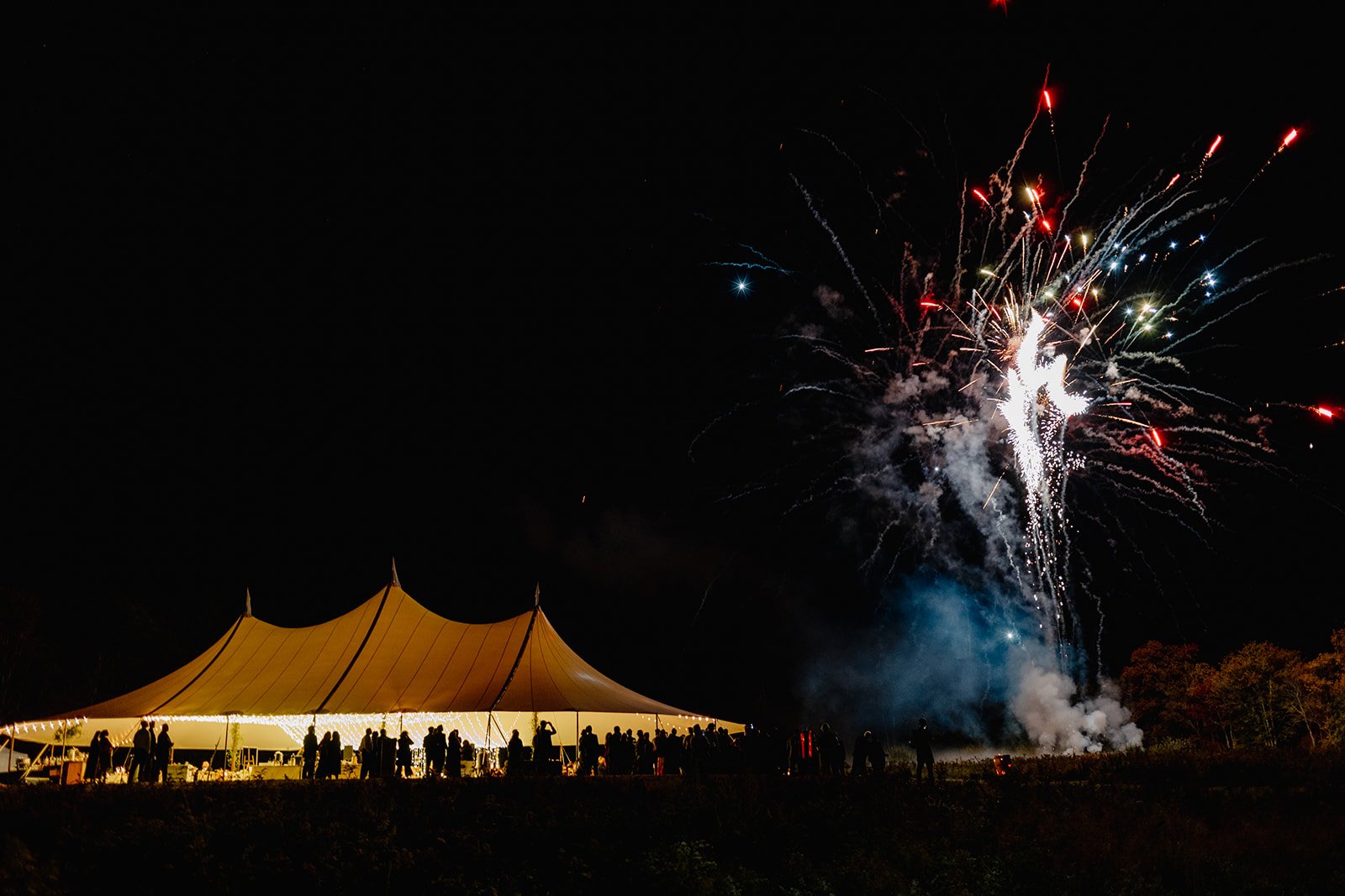 Nighttime scene with fireworks lighting up the sky and a large illuminated tent with people gathered underneath.