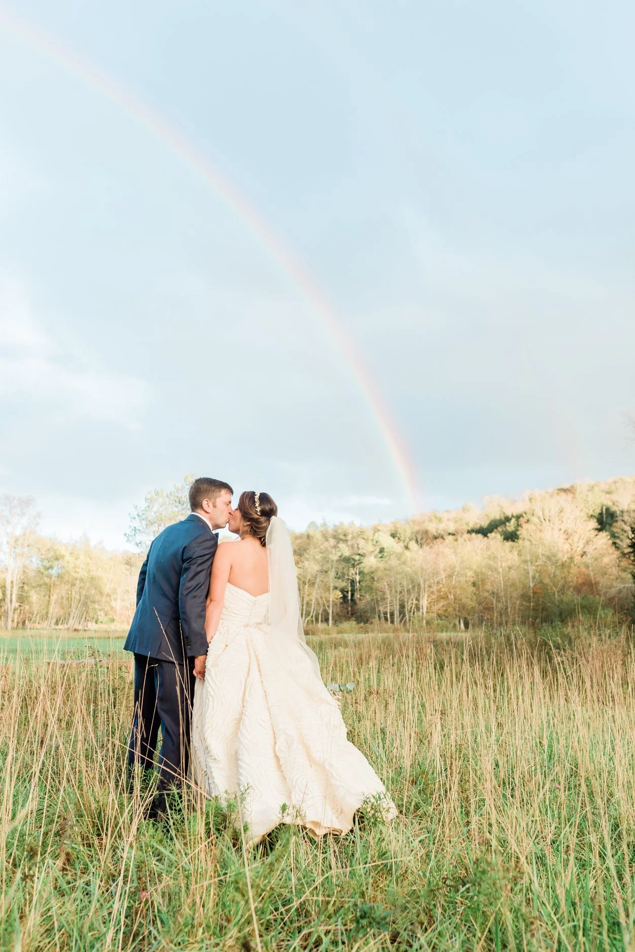 A bride and groom sharing a kiss outdoors in a field with tall grass, with a rainbow in the sky behind them.