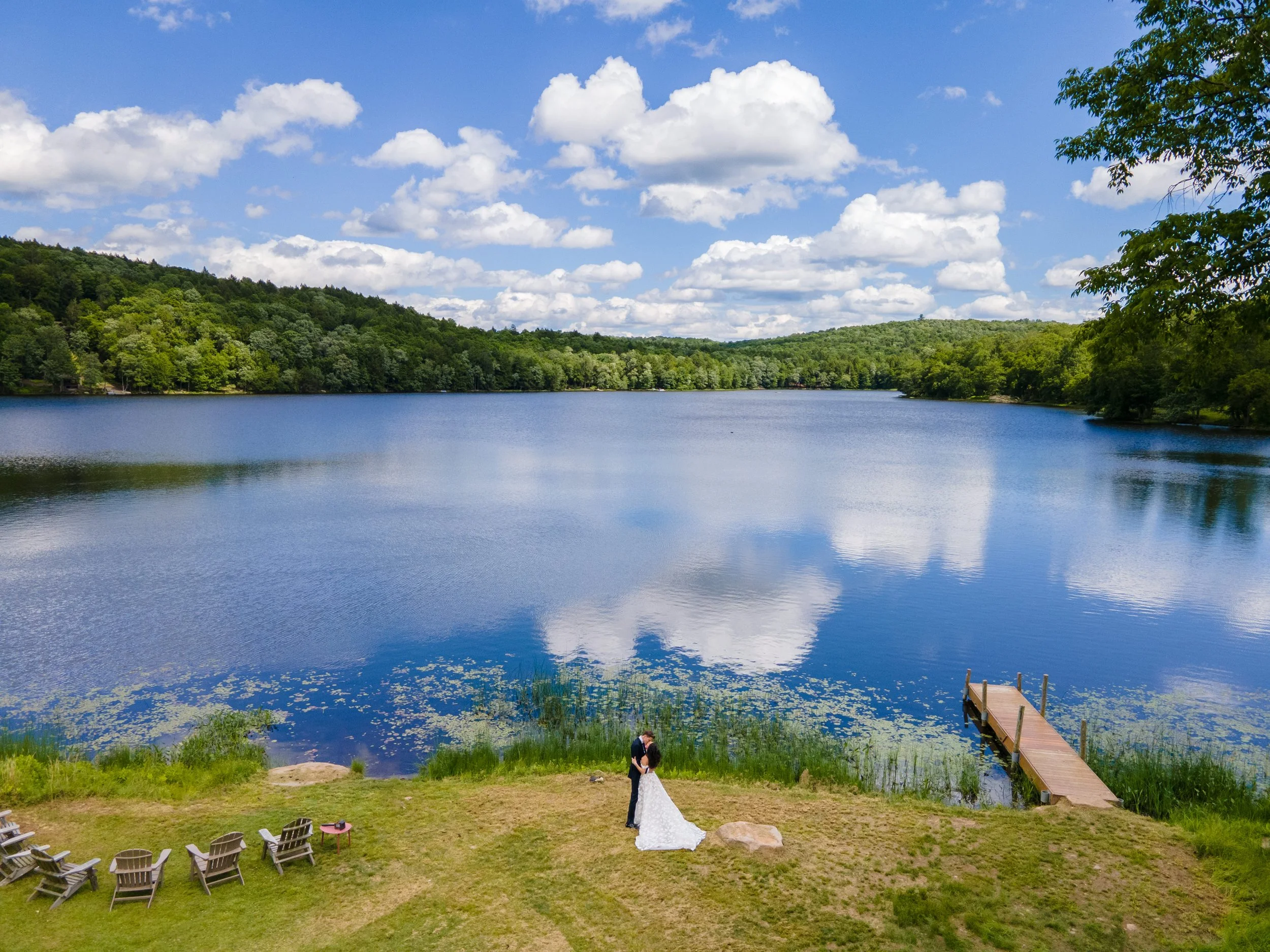 A bride and groom standing together on a grassy area near a lake, with several Adirondack chairs and a small dock in the background, surrounded by green trees and under a partly cloudy sky.