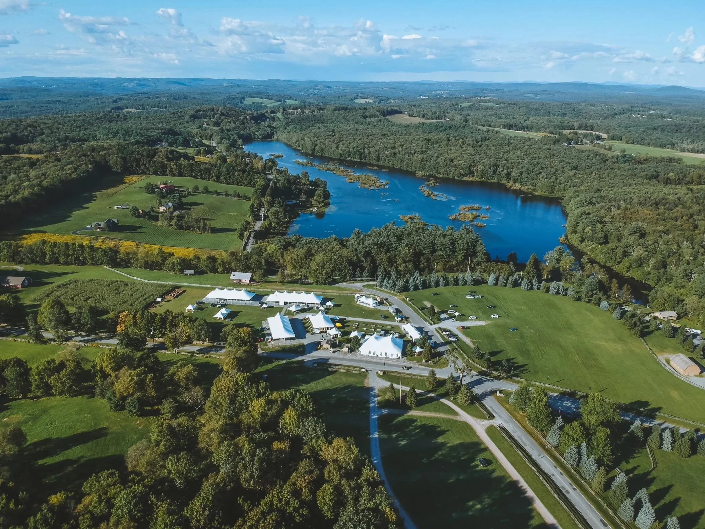 Aerial view of a large outdoor event with white tents set up on a green lawn next to a winding river, surrounded by trees and fields.