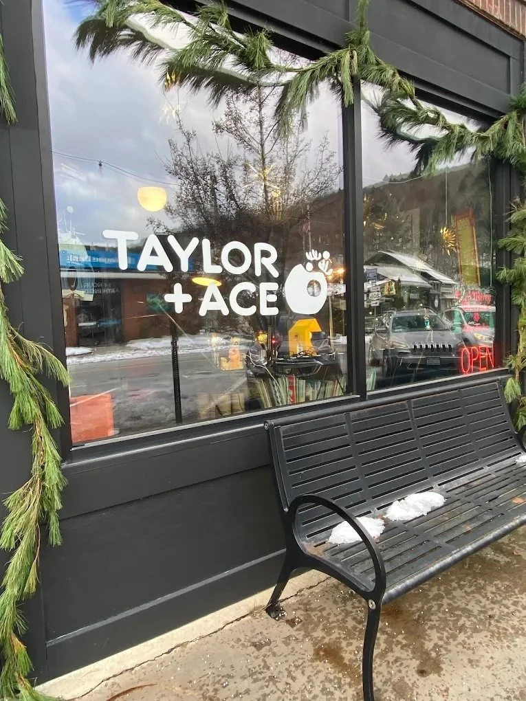 Store window with holiday greenery, a black bench with snow patches, and a sign that reads 'TAYLOR + ACE' with a decorative reindeer and ornament icon, illuminated 'OPEN' sign in red.