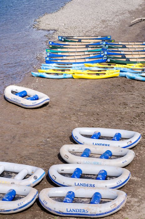 Multiple blue and yellow inflatable kayaks and white rubber boats are lined up on a sandy beach by the water.
