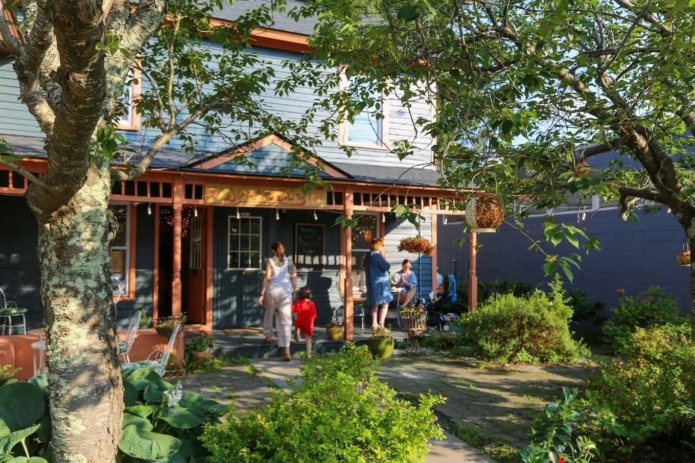 People sitting and standing on a porch outside a blue house with trees and plants surrounding them, during daytime.