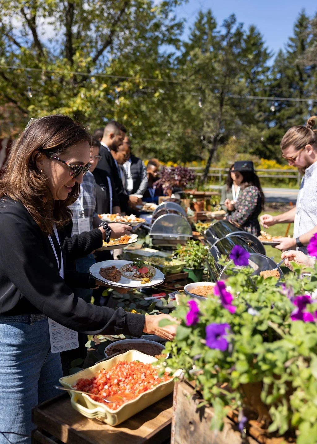 People are serving themselves food at an outdoor buffet table with various dishes, cakes, and salads, during a sunny day with trees in the background.