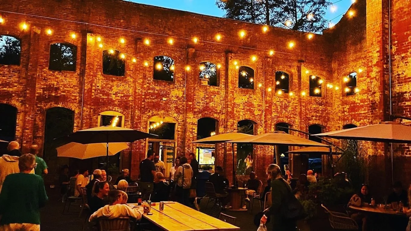 Outdoor patio of a brick building decorated with string lights, with people sitting and standing, some under umbrellas.