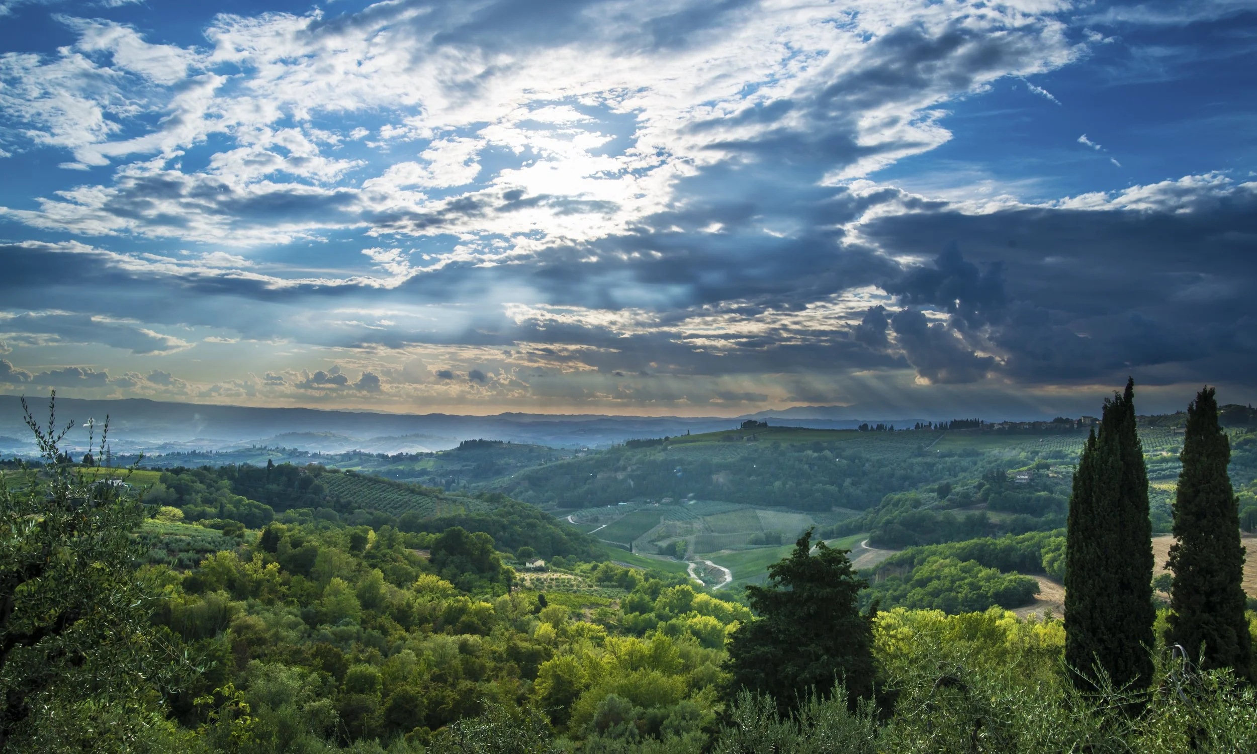 Scenic view of rolling green hills with trees, vineyards, and a winding road under a partly cloudy sky with the sun breaking through clouds