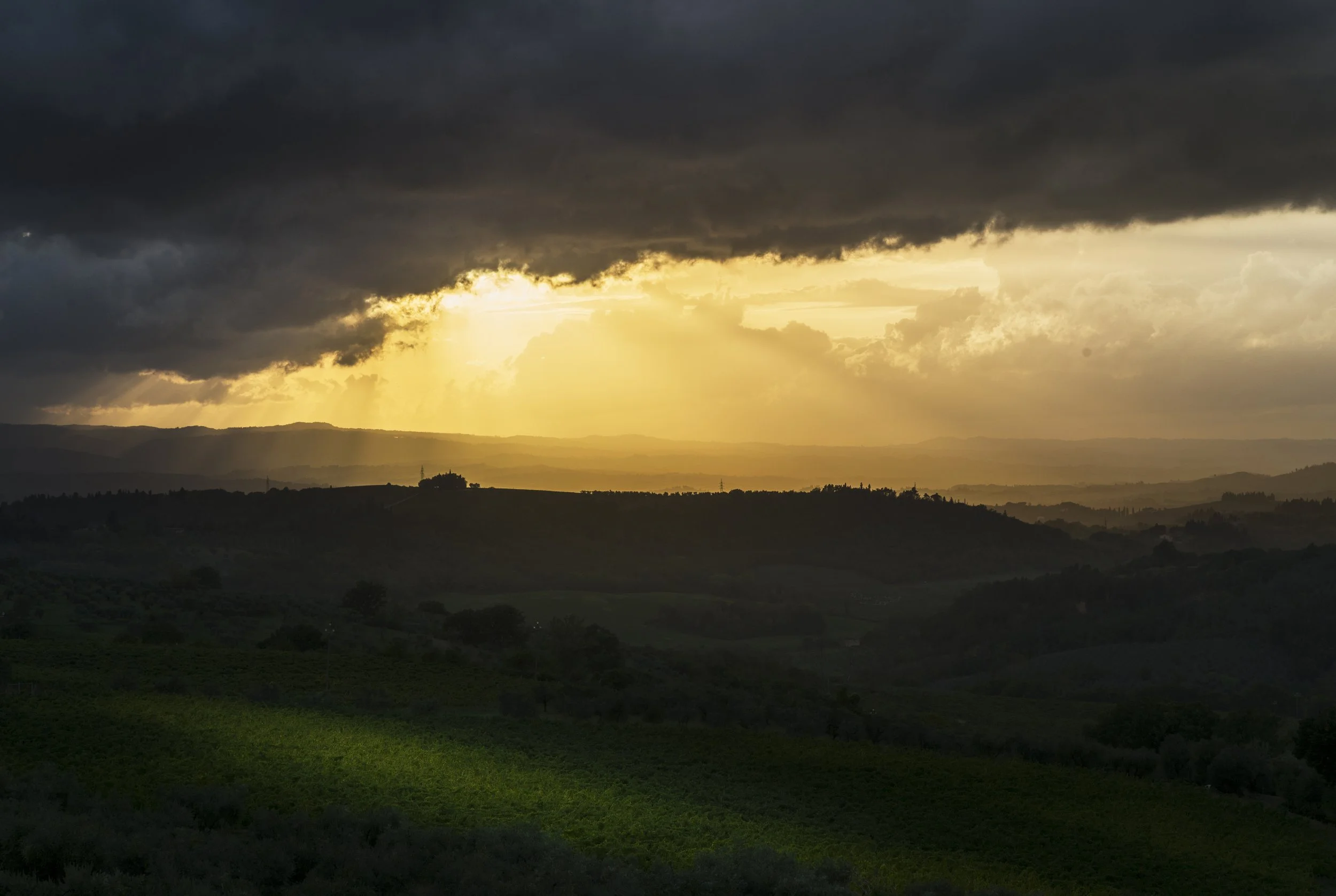 Dark storm clouds over rolling green hills as the sun sets, casting a golden glow through the clouds.