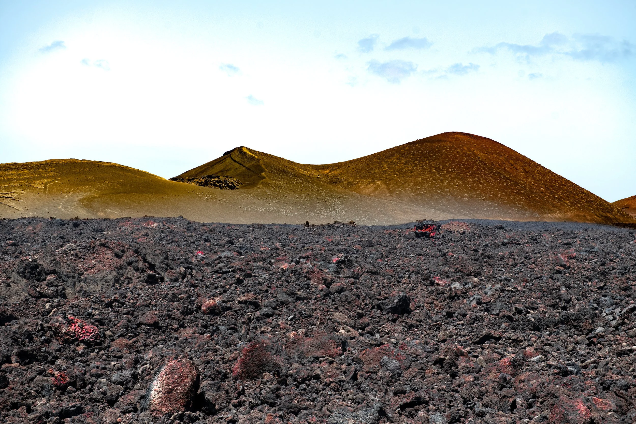 A landscape with dark, rough volcanic rocks in the foreground and smooth, rounded hills with sparse vegetation in the background under a cloudy sky.