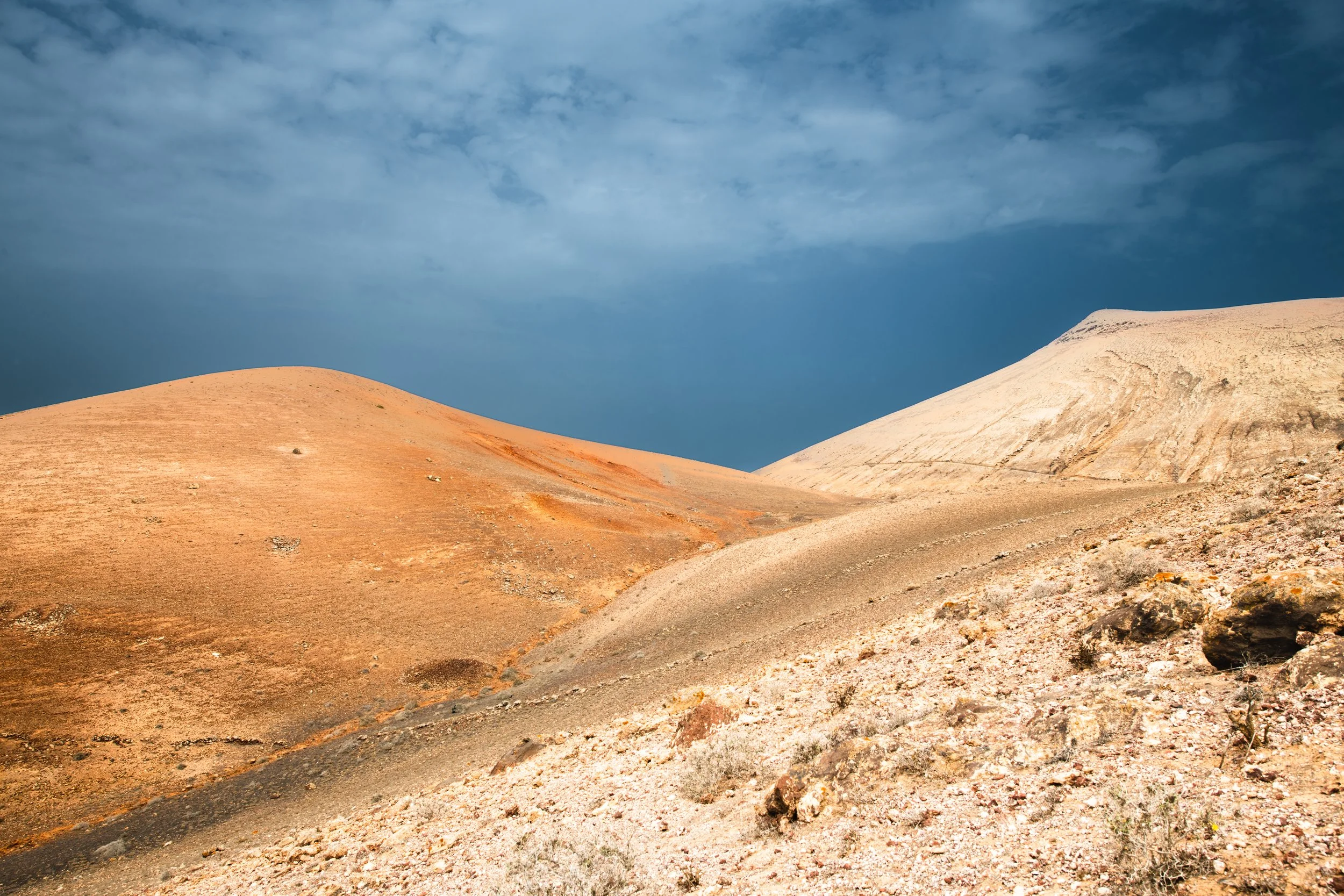 Desert landscape with orange and beige rolling hills and a dark blue cloudy sky.