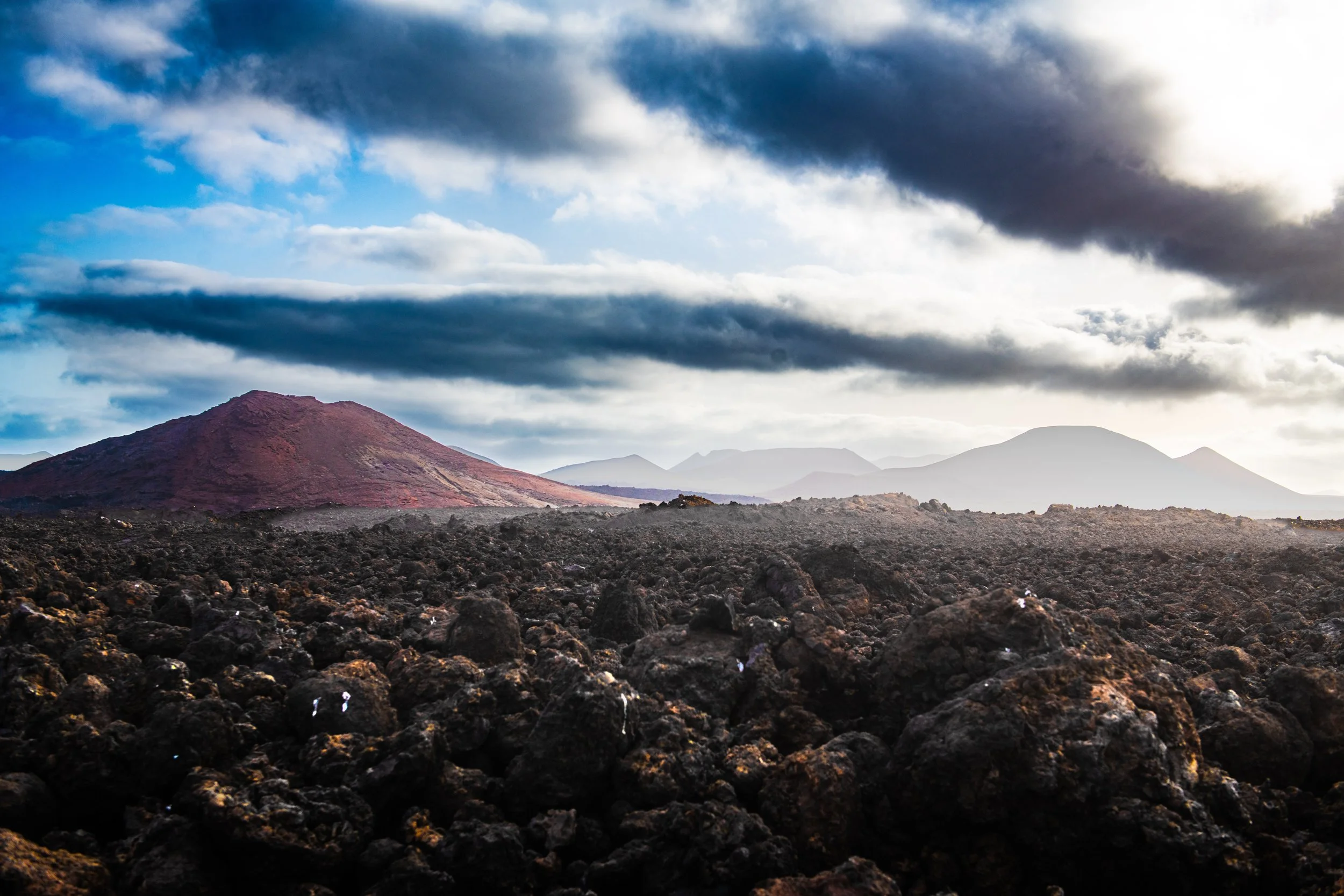 Volcanic landscape with dark lava rocks in the foreground and mountain peaks under a cloudy sky in the background.