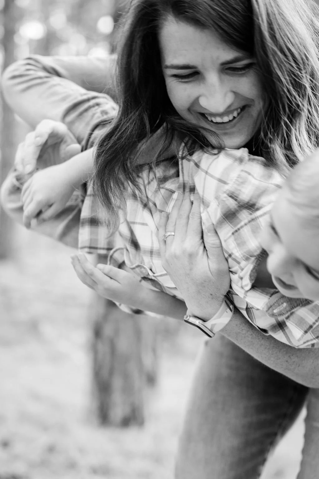 A woman with long hair and a smile, engaging in a playful hug with a child outdoors. The woman is wearing a plaid shirt, and the child's hands are visible.