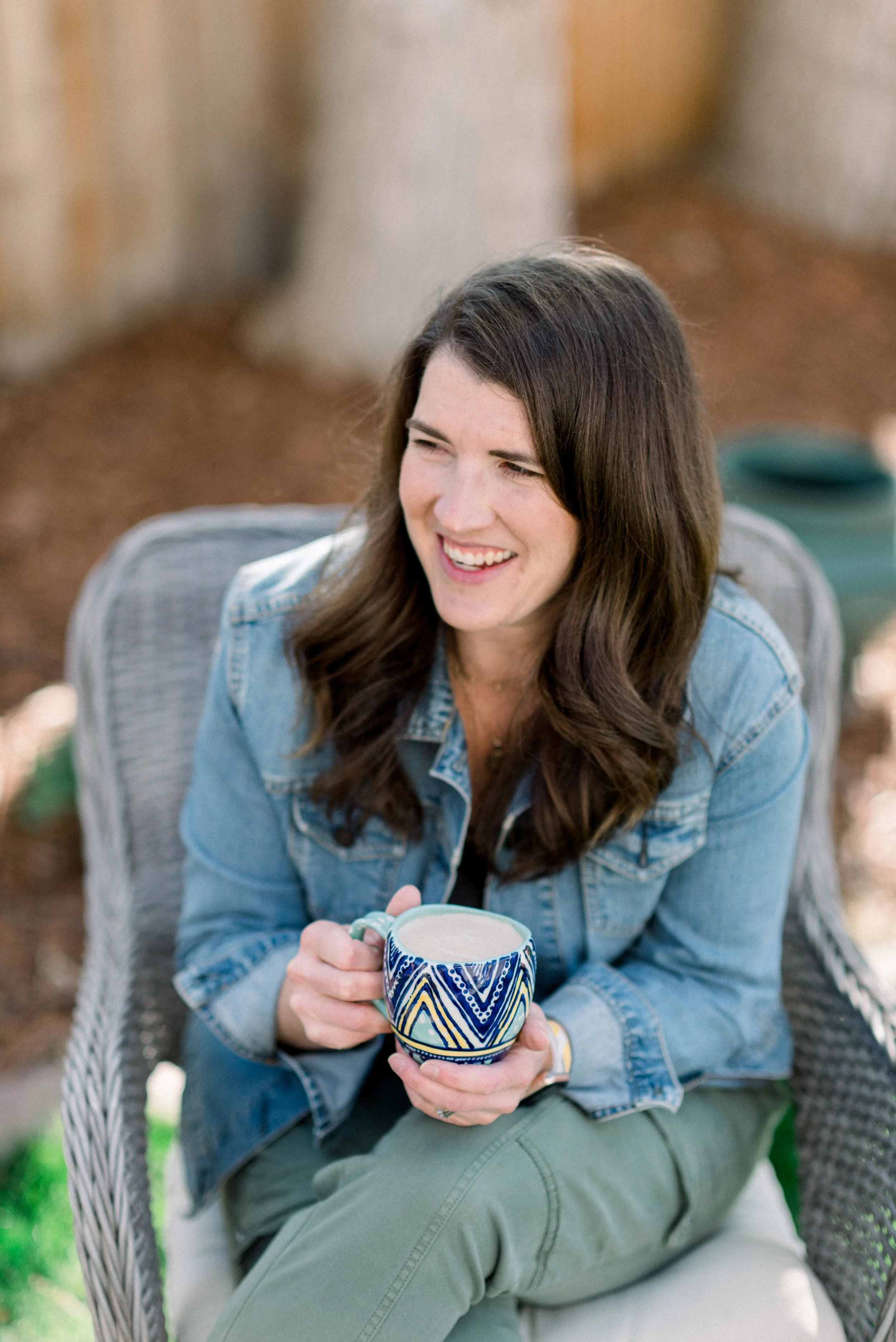 A woman sitting outdoors on a wicker chair, smiling and holding a colorful ceramic mug filled with a light-colored beverage.