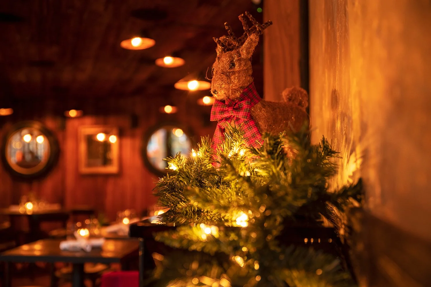 Decorative reindeer figurine with a red plaid bow on a Christmas garland with lights, against a warm, dimly lit wooden interior background.