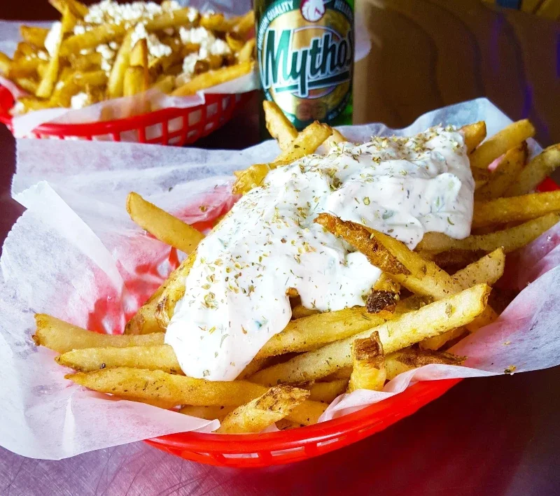 French fries topped with creamy white sauce and herbs in a red basket lined with paper, with a drink and another basket of fries in the background.