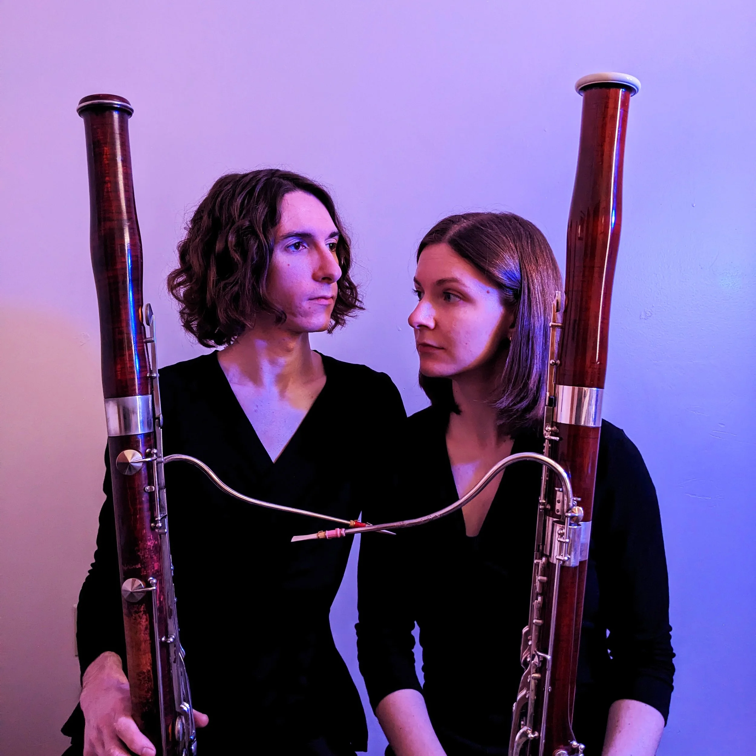 Bridge Bassoon duo headshot with muted blue-purple lighting. Andy Sledge and Dana Brink seated, facing past each other with neutral expressions. Both people are holding bassoons vertically.